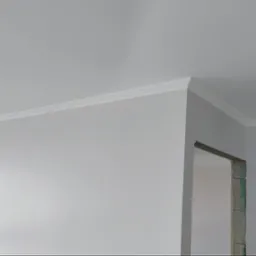 Wnętrze po remoncie: świeżo pomalowana ściana i sufit w odcieniach bieli, z widoczną dekoracyjną listwą przysufitową i surowym otworem drzwiowym w tle.