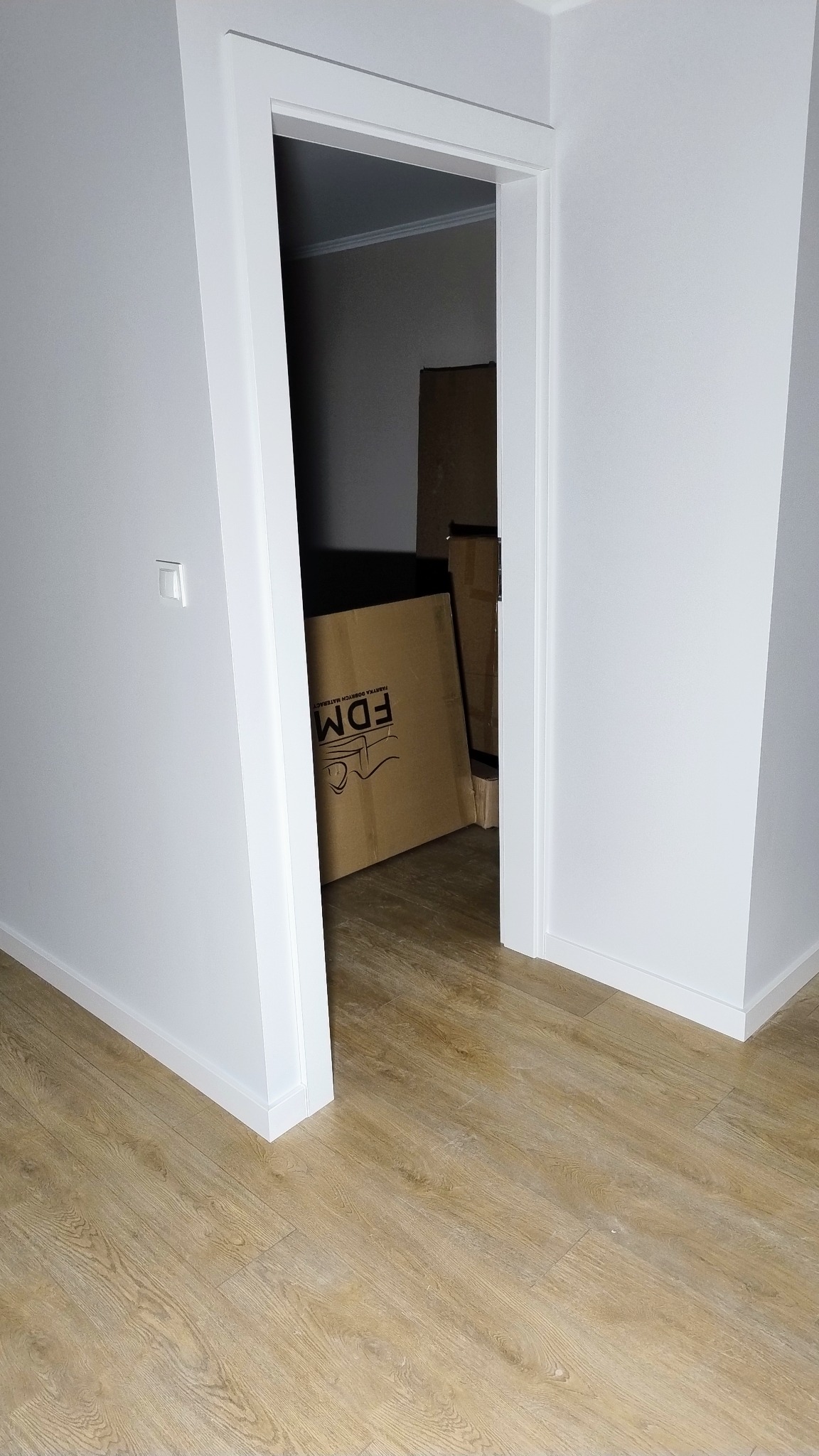 Pomalowane na biało ściany i ościeżnica drzwiowa w nowym mieszkaniu, widoczna podłoga z paneli oraz kartony w tle.