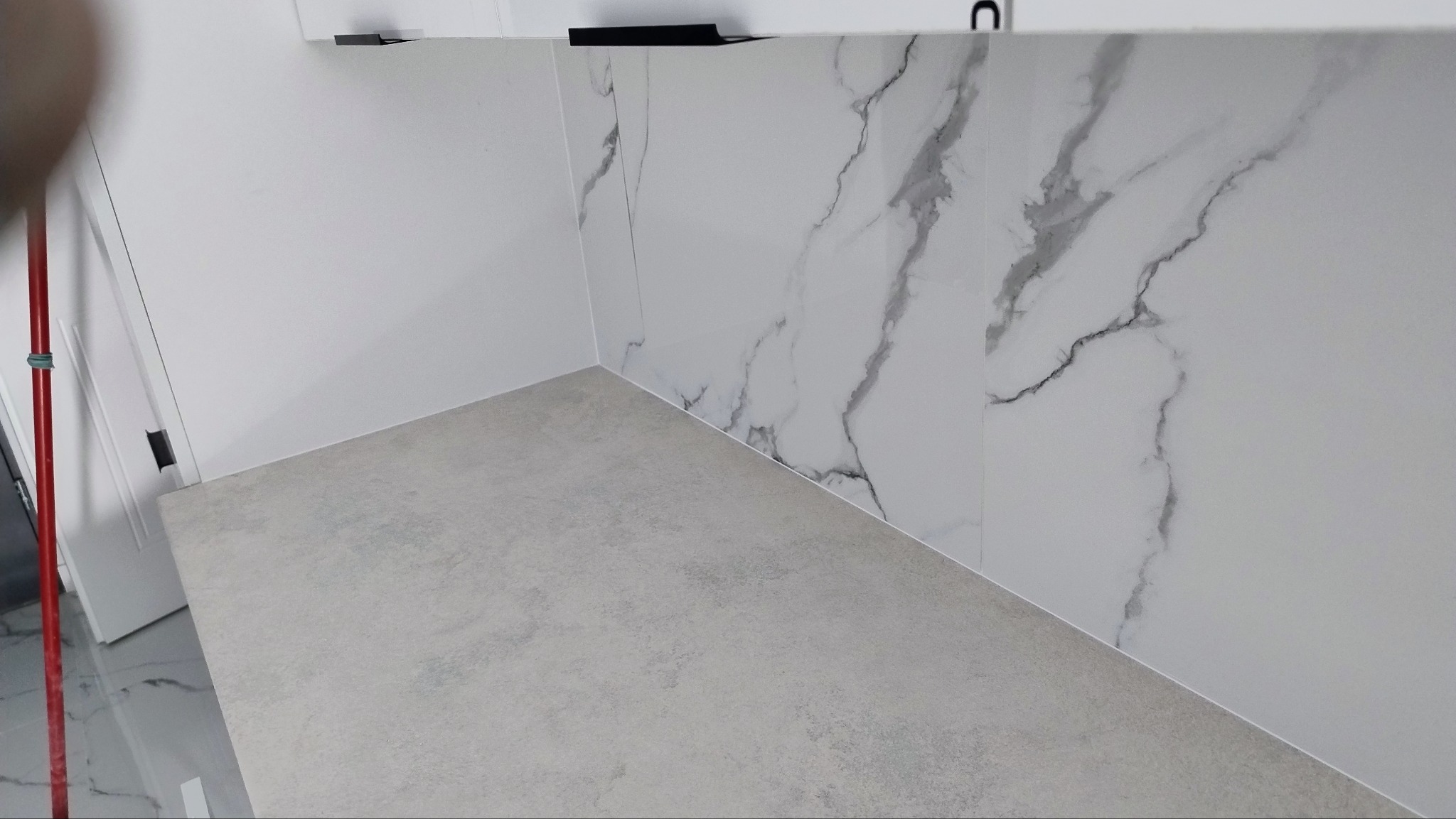 Wykończona ściana z białych płytek imitujących marmur i szara podłoga w rogu pomieszczenia, widoczny fragment czerwonego kija do mopa.