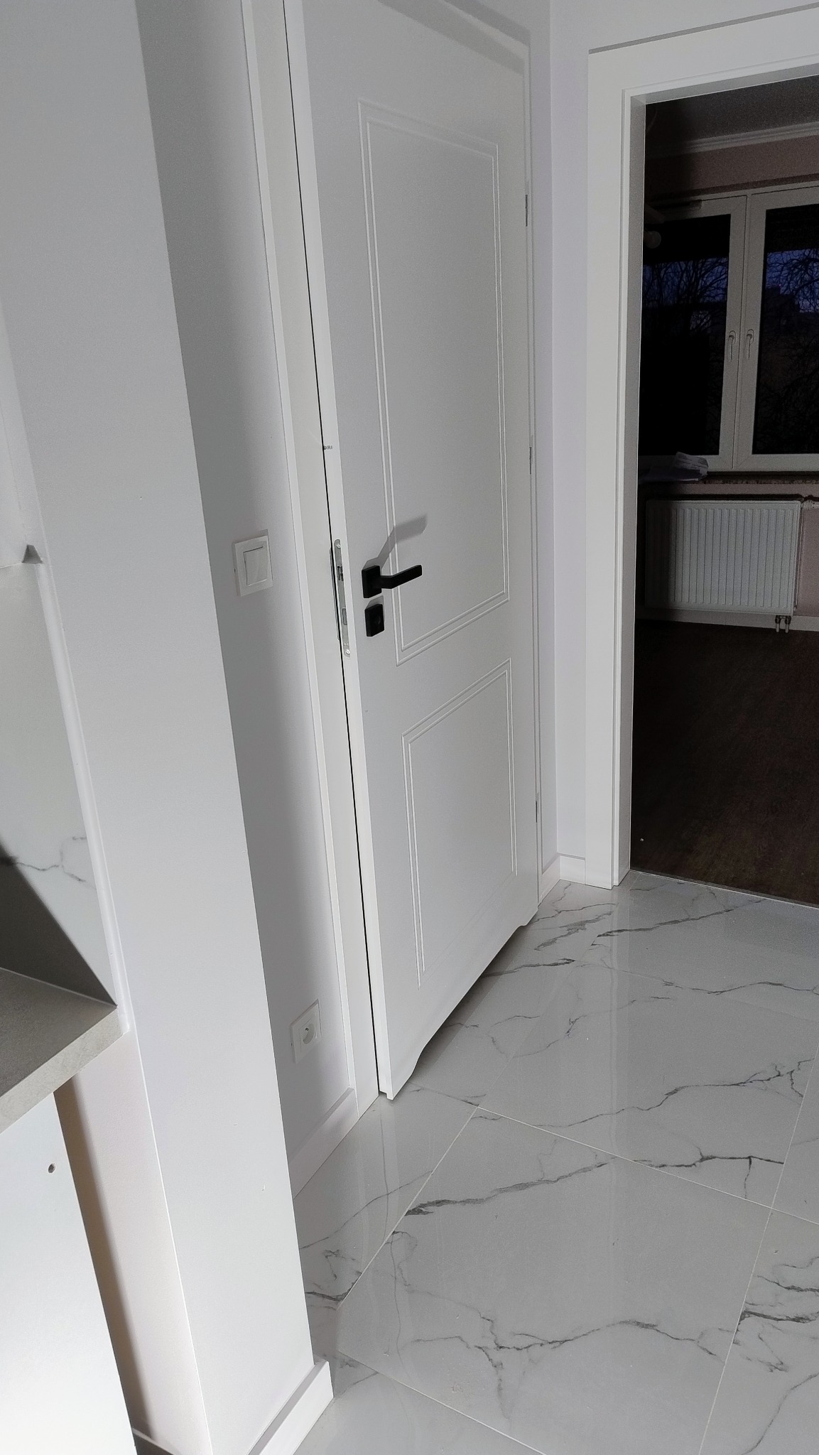 Białe, panelowe drzwi wewnętrzne z czarną klamką na tle białej ściany, widoczne płytki podłogowe imitujące marmur i fragment pomieszczenia za drzwiami z grzejnikiem i oknem.