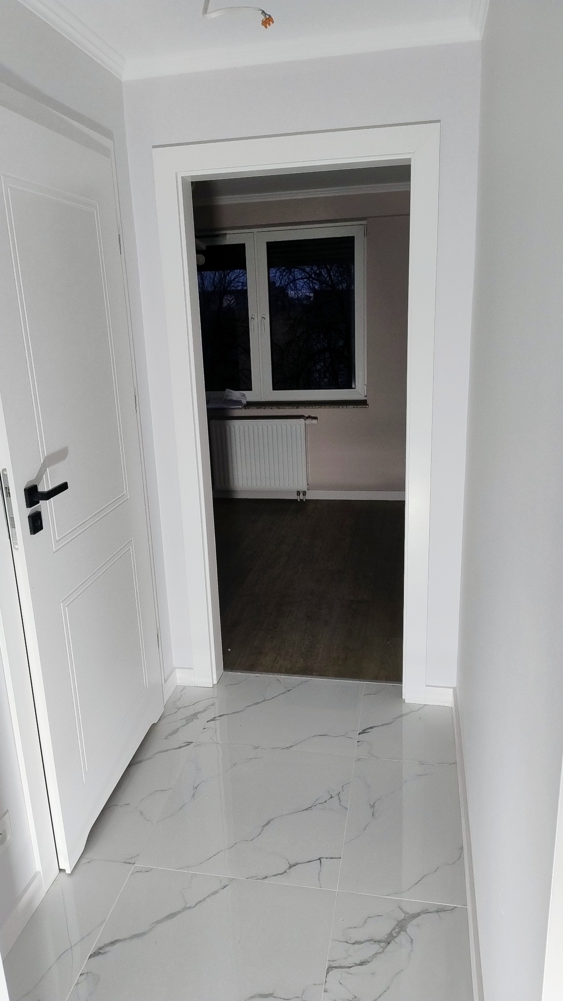 Białe, błyszczące płytki podłogowe z szarymi żyłkami w korytarzu, widok na pokój z ciemną podłogą przez białe drzwi i ościeżnicę.