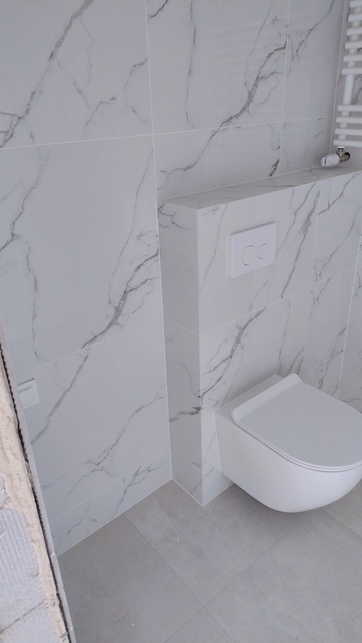 Nowoczesna toaleta podwieszana z białymi płytkami imitującymi marmur na ścianach i szarą podłogą, widoczny fragment grzejnika drabinkowego i niedokończone wykończenie po lewej stronie.