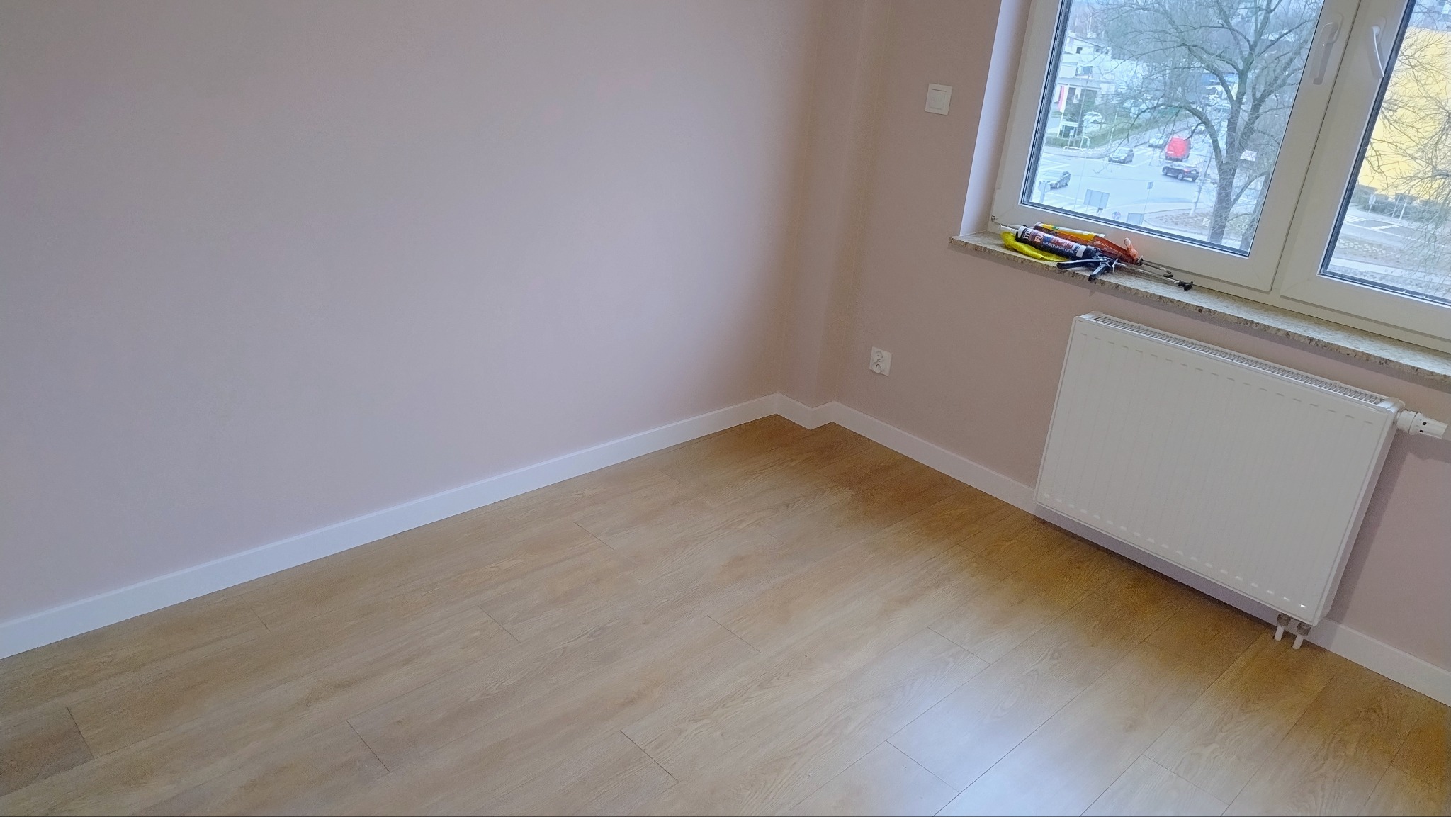 Wnętrze pokoju po remoncie: świeżo pomalowane ściany na delikatny róż, jasne panele podłogowe, białe listwy przypodłogowe i grzejnik pod oknem z narzędziami na parapecie.