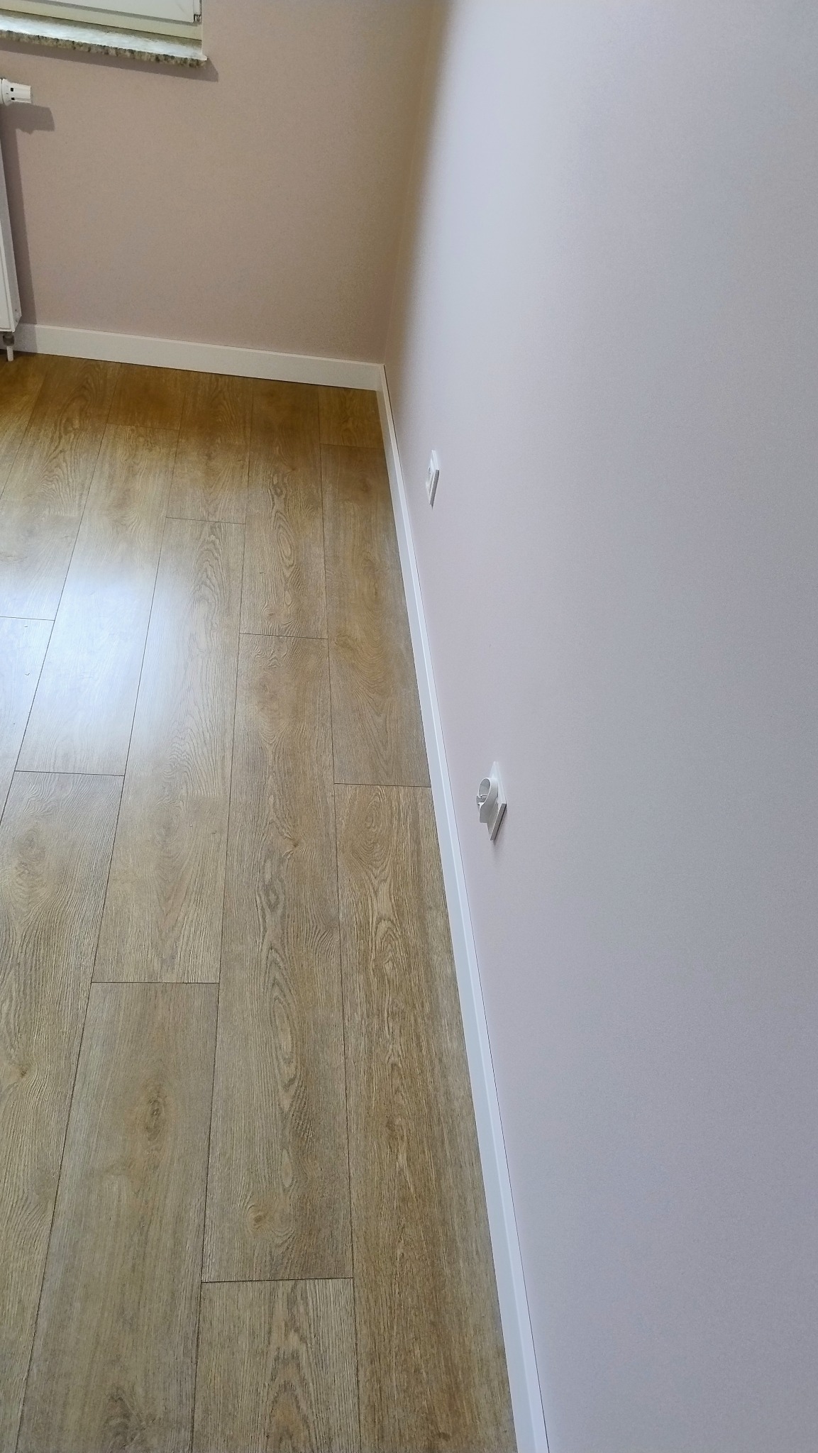 Jasne panele podłogowe ułożone wzdłuż ściany pomalowanej na delikatny róż, z widoczną białą listwą przypodłogową i gniazdkami elektrycznymi.