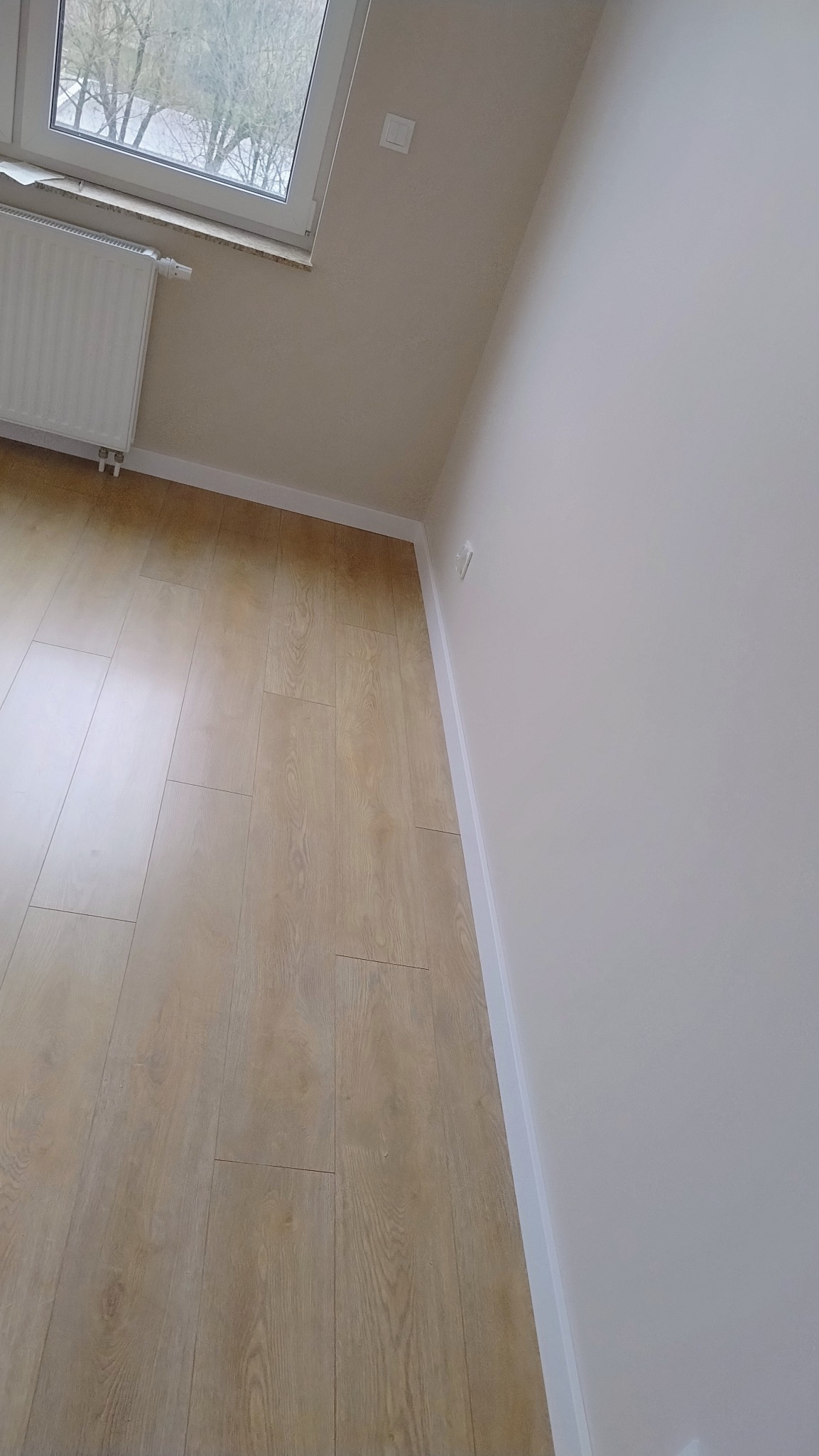 Świeżo wyremontowane wnętrze z panelami podłogowymi w odcieniach jasnego drewna, pomalowane na biało ściany z listwami przypodłogowymi, okno z grzejnikiem oraz gniazdko elektryczne.