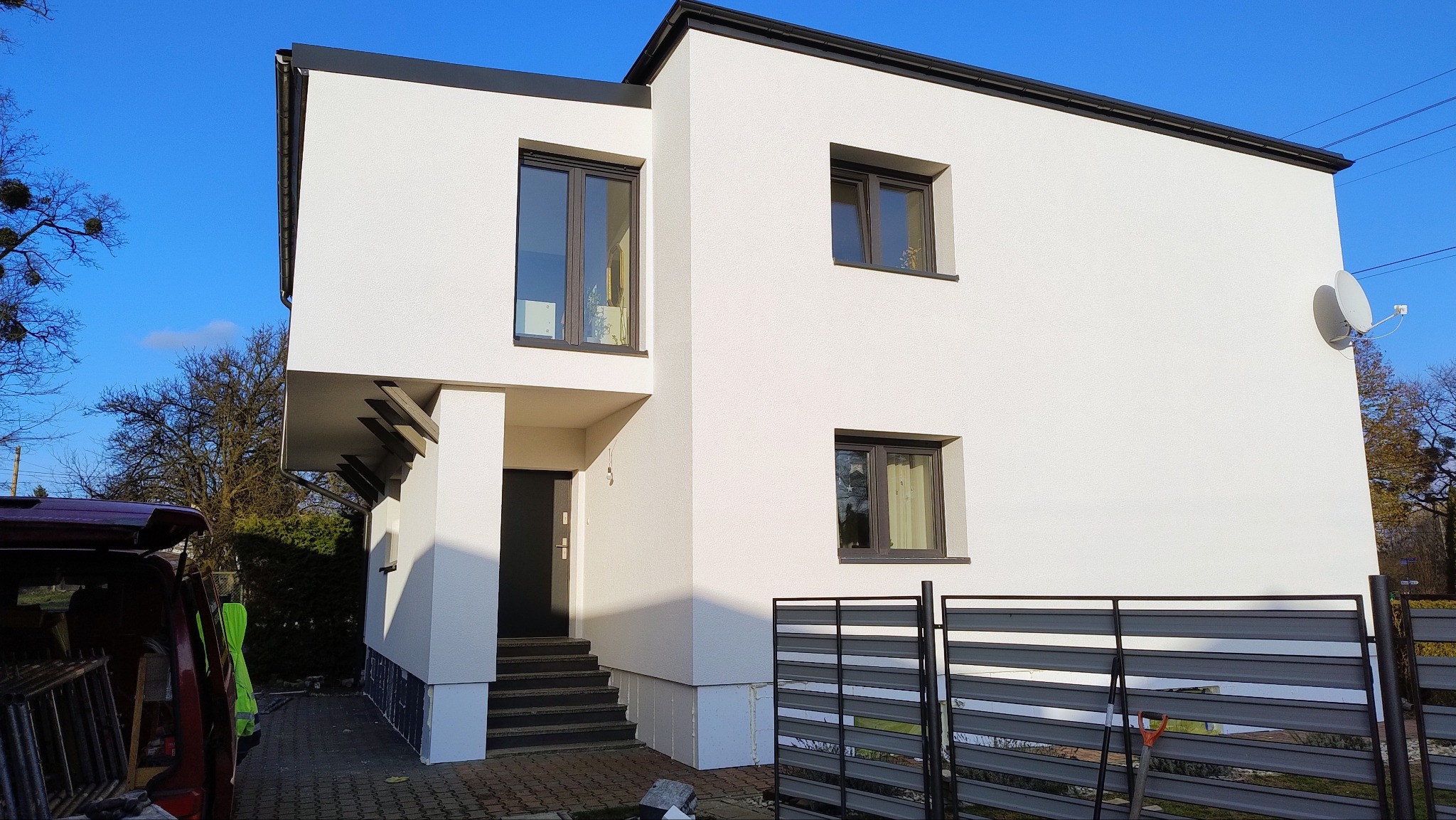 Dwupiętrowy dom z białą elewacją i ciemnymi oknami, widoczna antena satelitarna i fragment samochodu dostawczego z narzędziami.