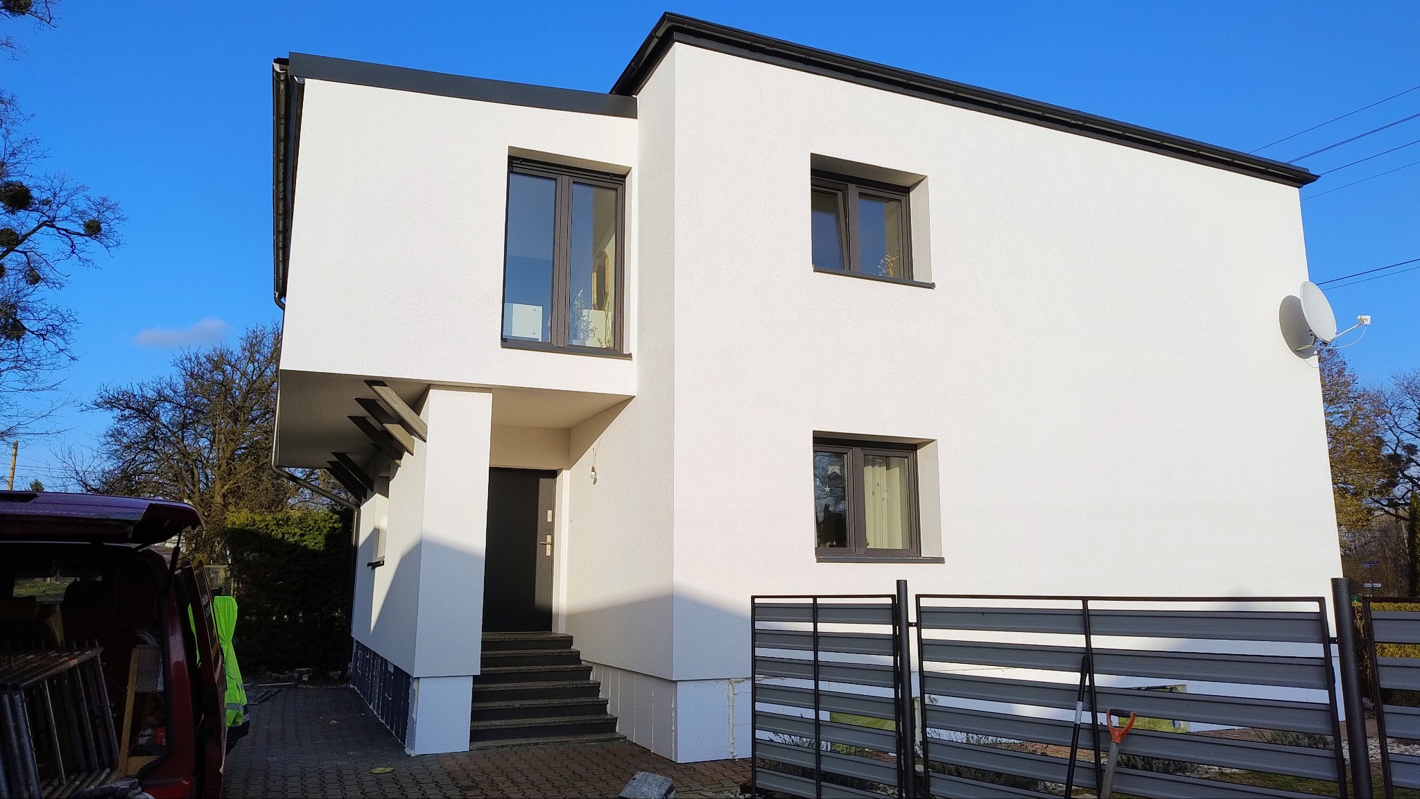 Nowoczesny dom jednorodzinny z białą elewacją i ciemnymi oknami, widoczny fragment samochodu z narzędziami sugerujący prace wykończeniowe.