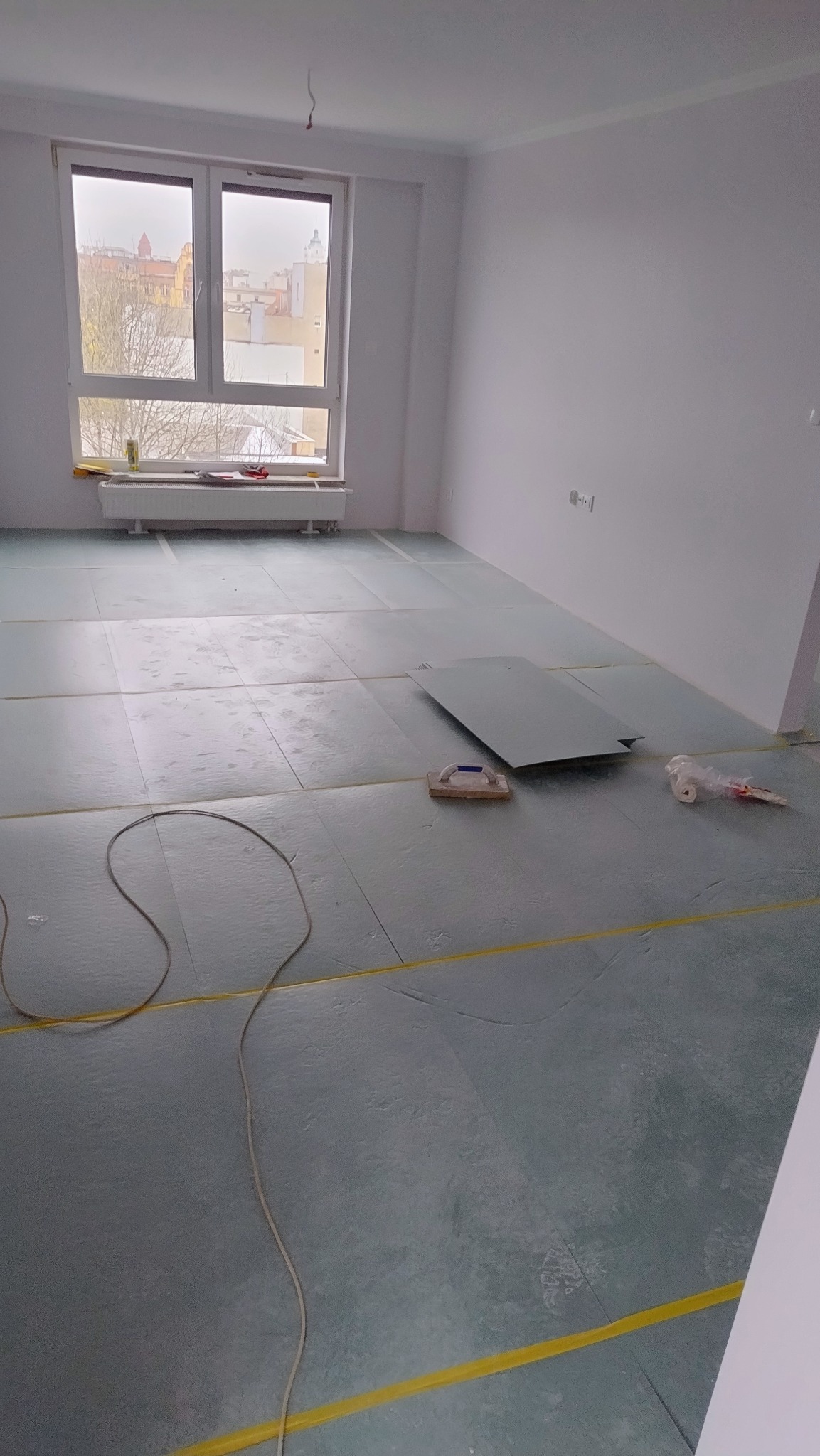 Przygotowanie podłoża pod panele: rozłożona pianka izolacyjna z żółtą taśmą, widoczny fragment płyty i narzędzie do docinania, surowe ściany i okno z widokiem na miasto.