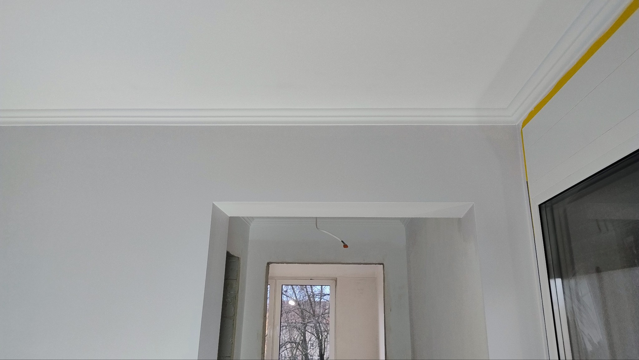 Szare ściany w trakcie remontu, widoczne listwy dekoracyjne przy suficie, żółta taśma malarska zabezpieczająca okno, oraz surowy otwór drzwiowy z wystającym kablem.