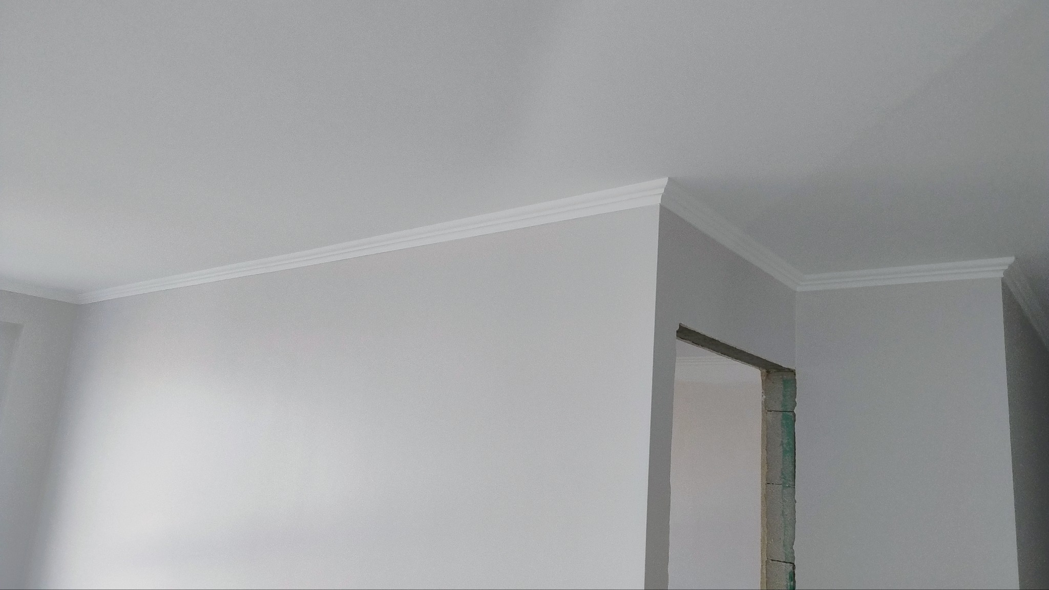 Wnętrze po remoncie: świeżo pomalowana ściana i sufit w odcieniach bieli, z widoczną dekoracyjną listwą przysufitową i surowym otworem drzwiowym w tle.