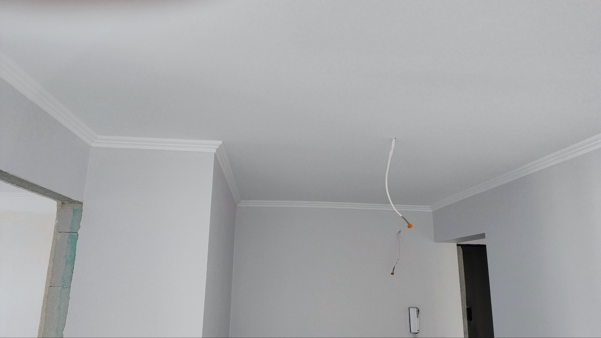 Świeżo pomalowane na szaro ściany i sufit z białymi listwami dekoracyjnymi przy suficie, widoczny wystający kabel elektryczny i fragment otworu drzwiowego w stanie surowym.