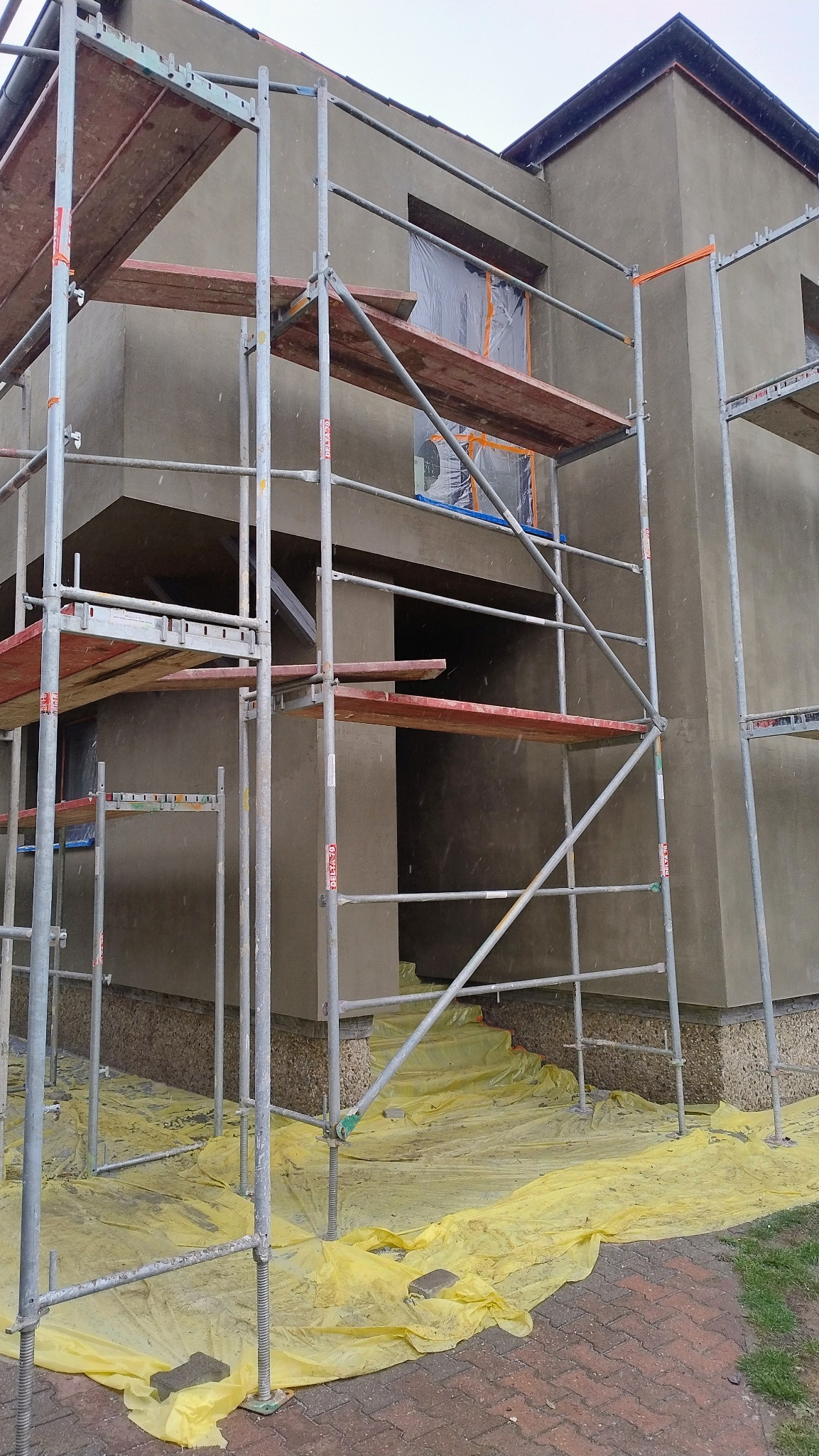 Budynek w trakcie nakładania tynku zewnętrznego, widoczne rusztowania, okna zabezpieczone folią, teren wokół budynku przykryty żółtą folią ochronną.
