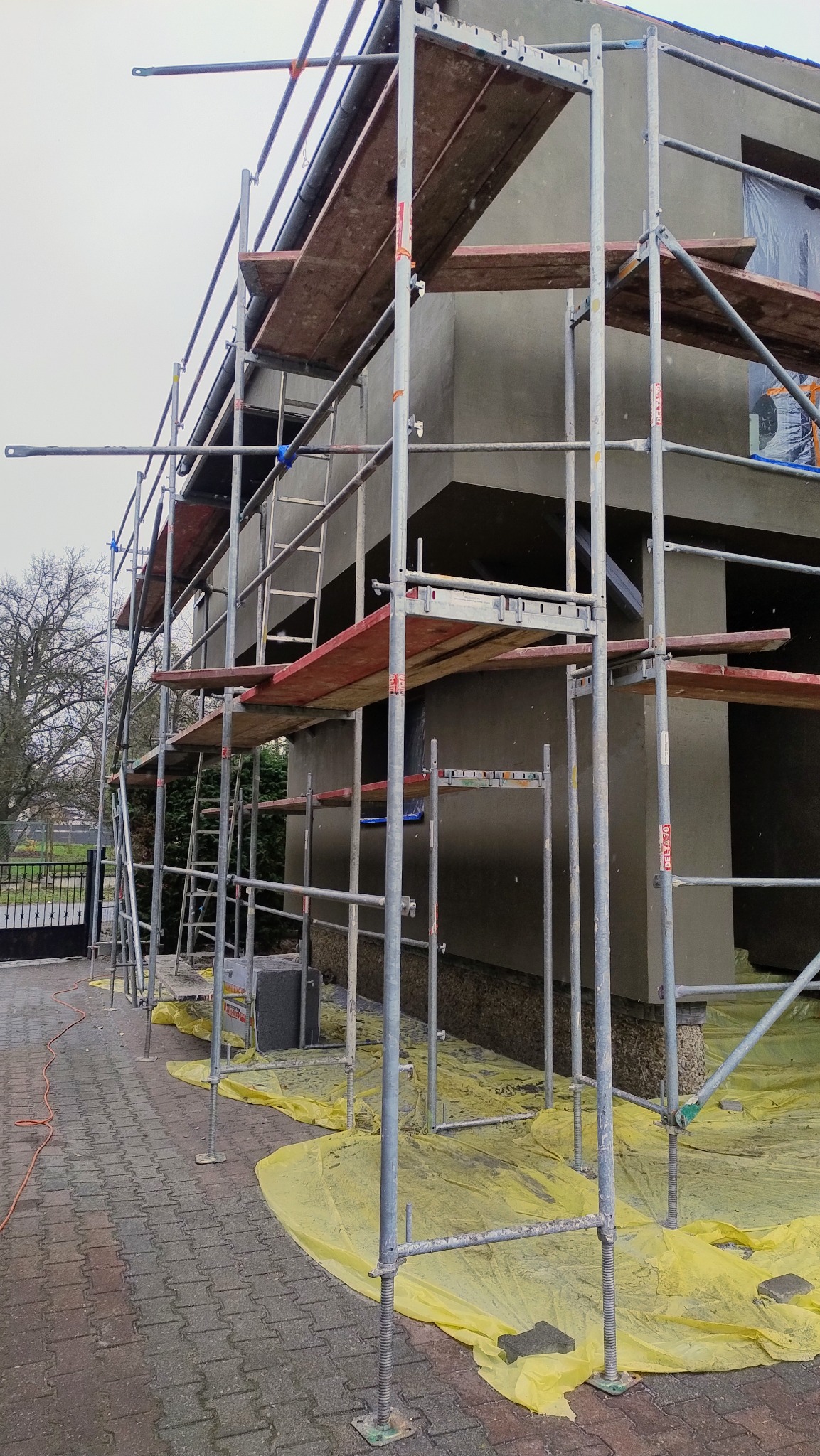 Dom w trakcie prac elewacyjnych, otoczony rusztowaniem i zabezpieczony folią, z widocznym fragmentem chodnika z kostki.
