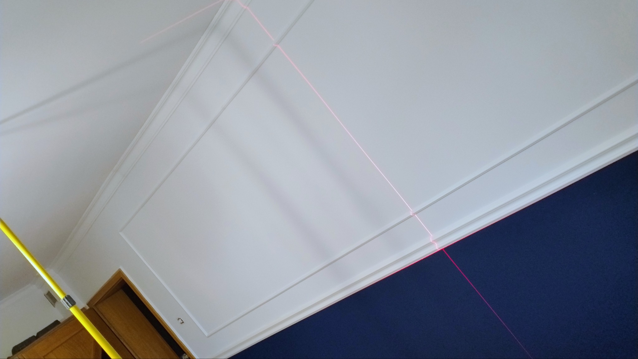 Biała ściana z dekoracyjnymi listwami i granatową ścianą w tle, wyznaczona liniami lasera, widoczny fragment żółtej poziomicy.