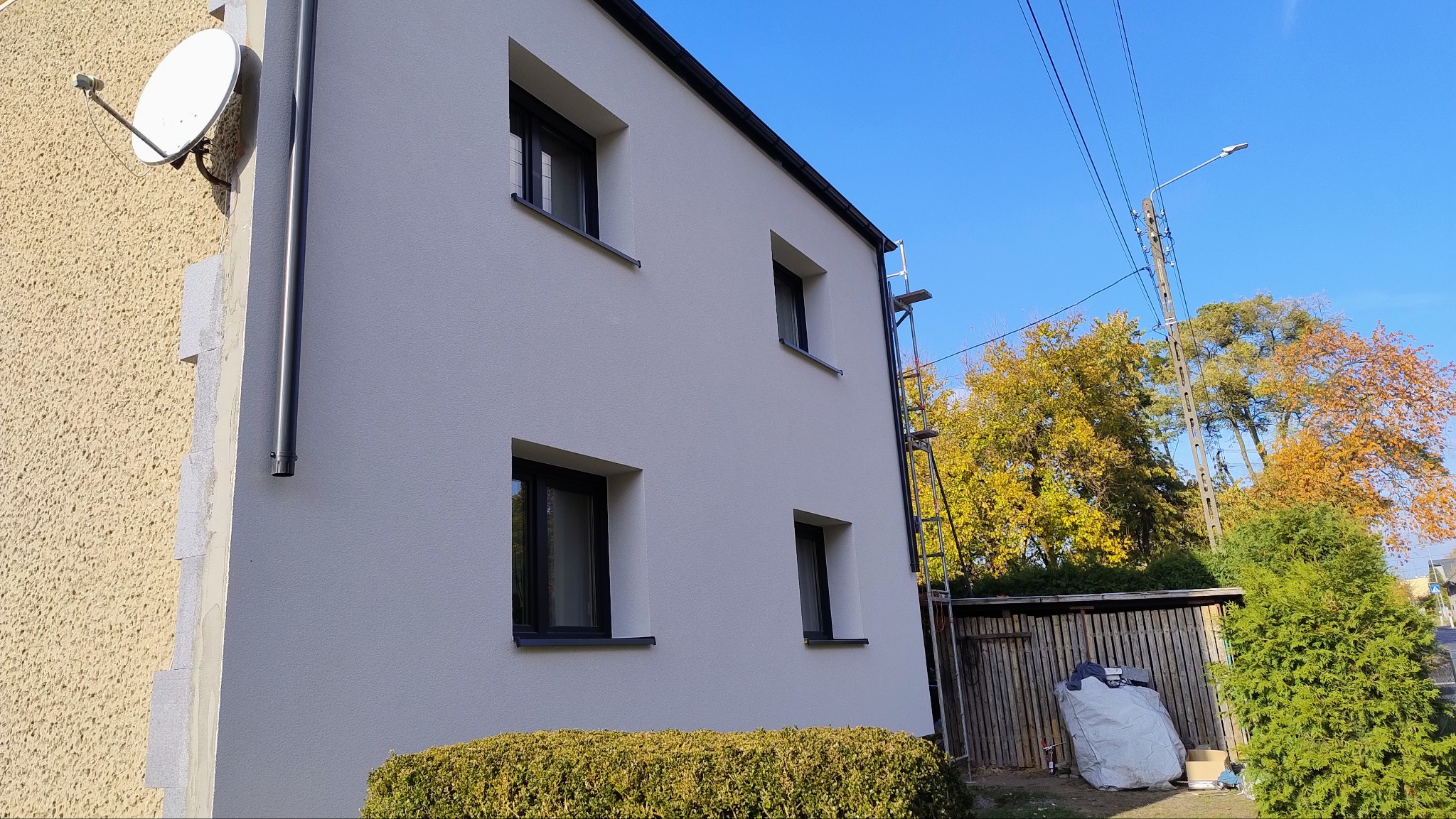 Fragment elewacji budynku po odnowieniu, z widoczną nową, białą strukturą, czarnymi ramami okiennymi i częścią elewacji w innym kolorze, z zamontowaną anteną satelitarną, drabina oparta o ścianę.