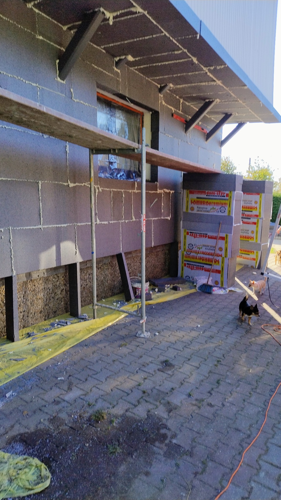 Budynek w trakcie prac związanych z izolacją termiczną ścian zewnętrznych, widoczne rusztowanie, płyty izolacyjne, materiały budowlane, dwa psy w pobliżu.