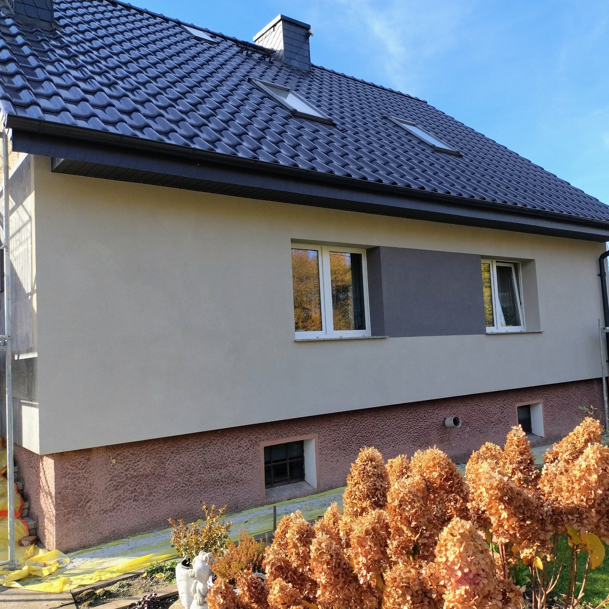 Wykończona elewacja domu jednorodzinnego z szarą wstawką, widoczne okna z odbiciem drzew, dachówka i fragment rusztowania. U dołu brązowa podmurówka i roślinność.