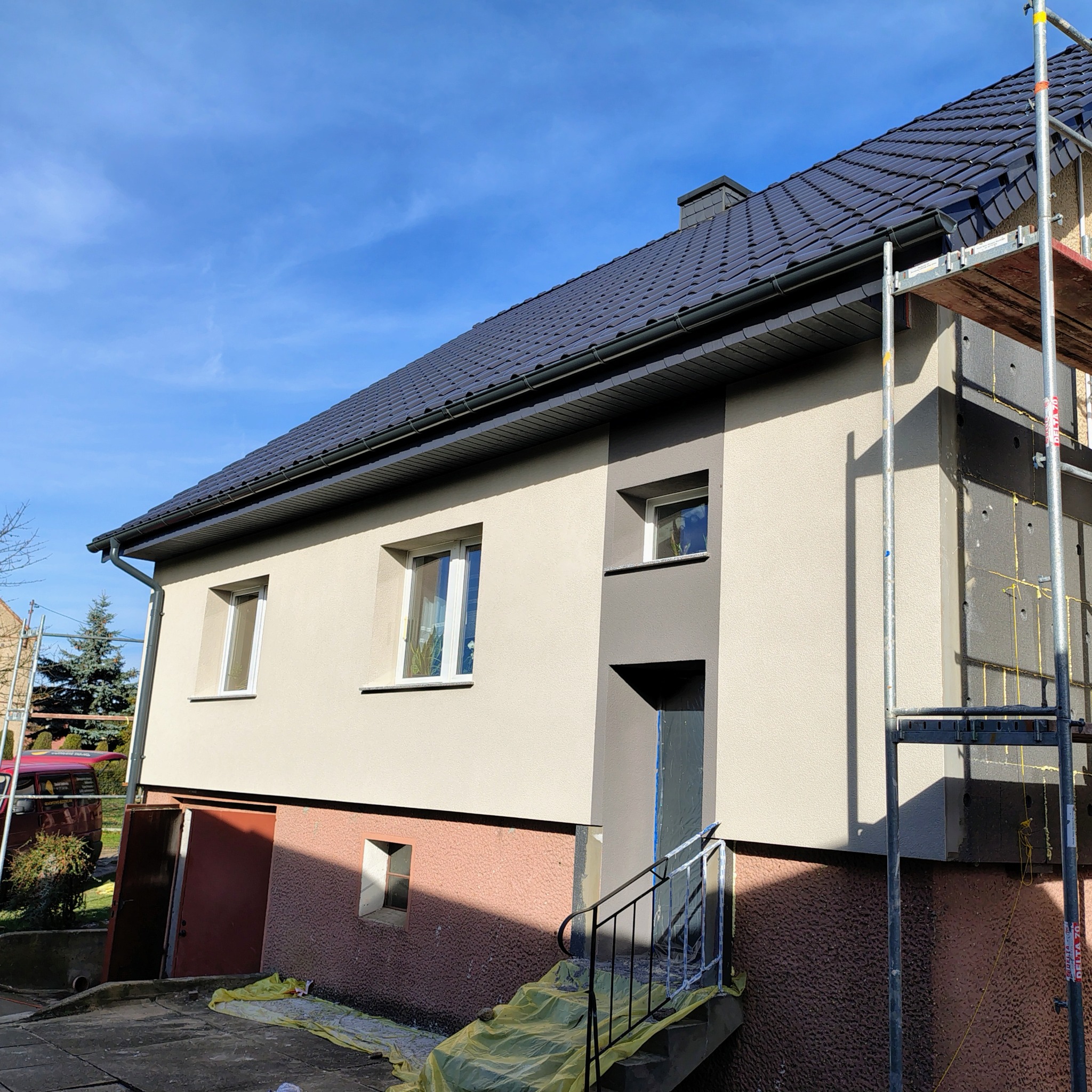 Dom po remoncie elewacji z rusztowaniem. Część ściany ocieplona styropianem, reszta pomalowana na beżowo z szarymi akcentami. Nowy dach z ciemnej dachówki.