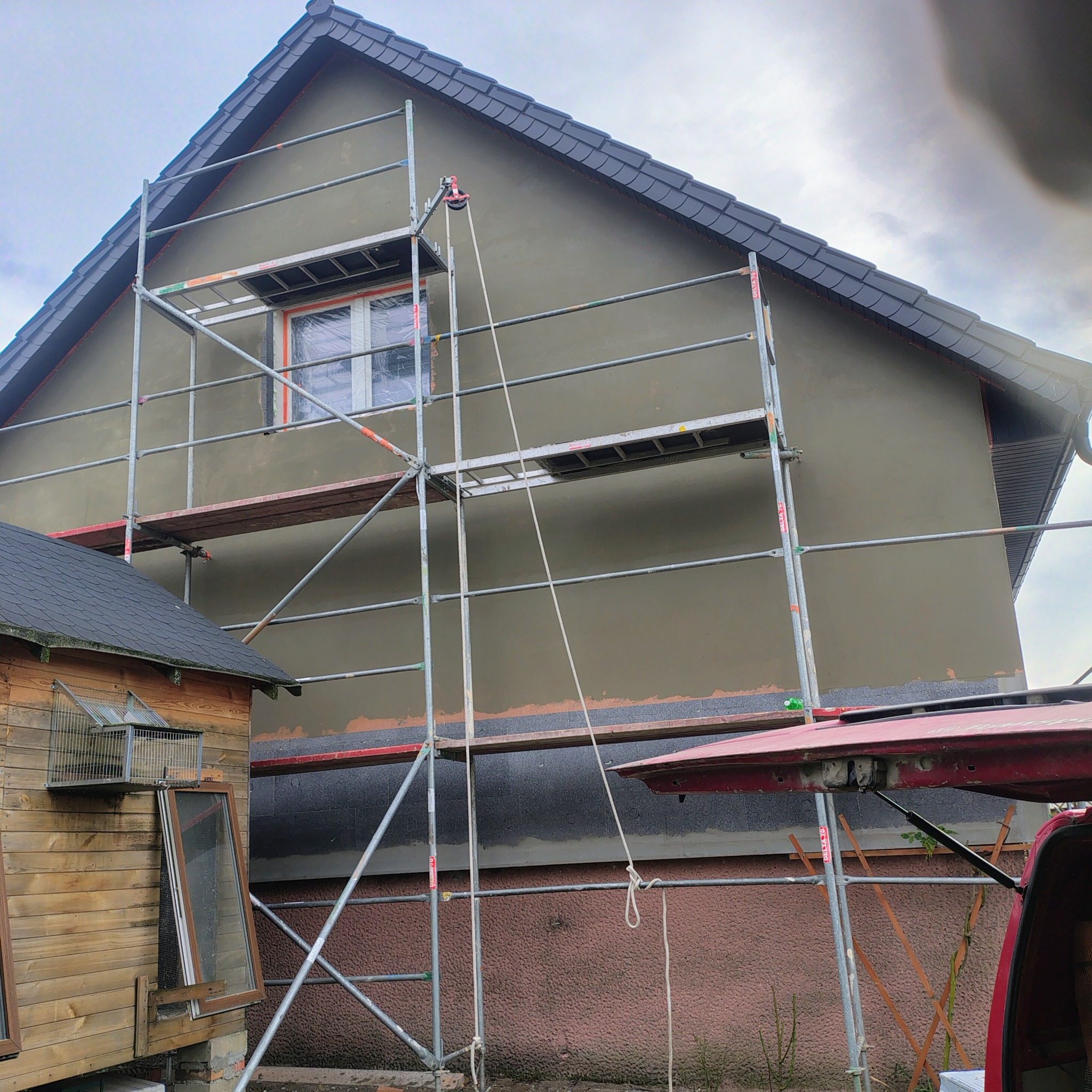 Dom w trakcie nakładania tynku elewacyjnego, widoczne rusztowania i fragmenty ocieplenia. Prace na wysokościach, szara elewacja, dach z ciemnej dachówki.