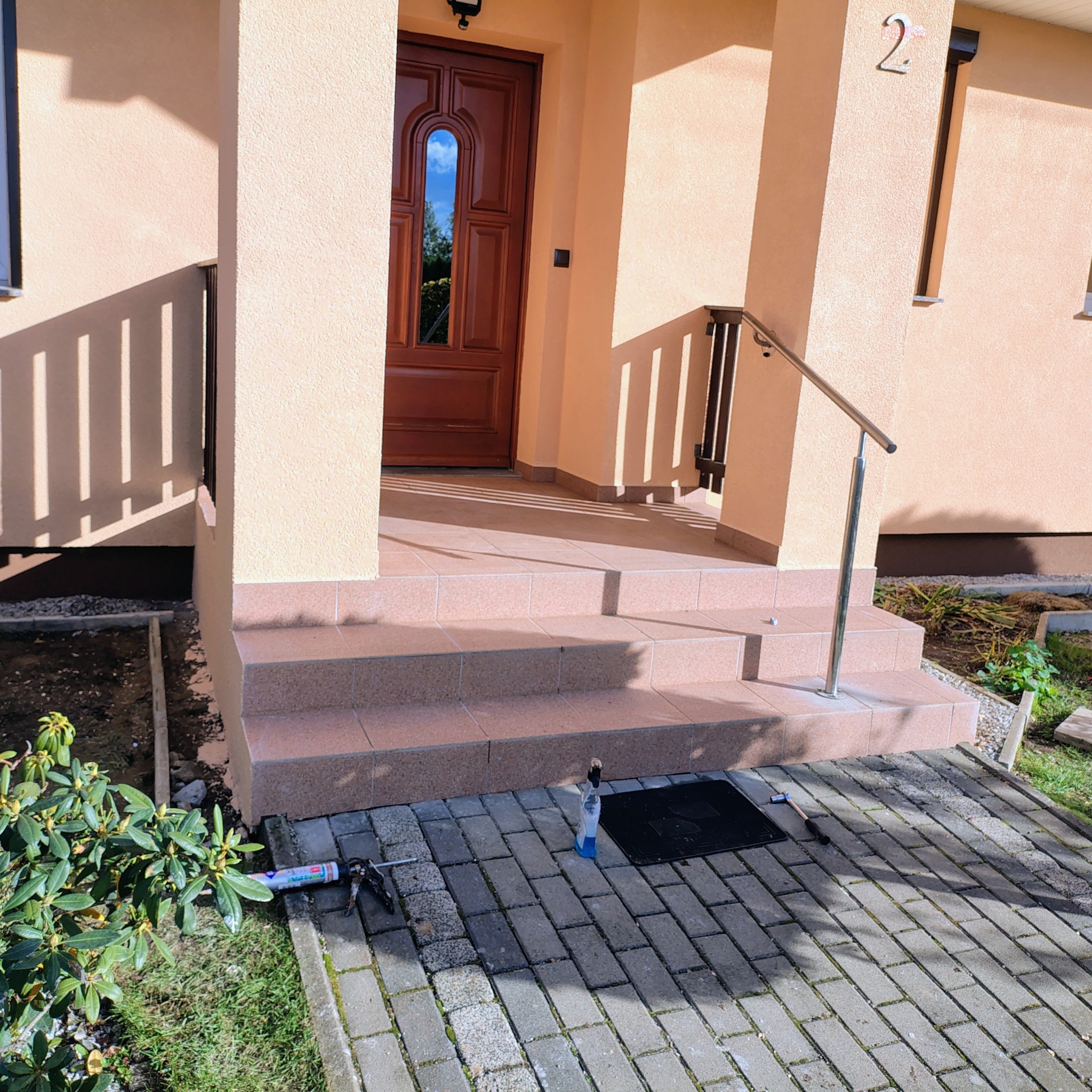 Wejście do domu z brązowymi drzwiami i schodami wyłożonymi płytkami. Widoczne narzędzia i materiały budowlane sugerują prace wykończeniowe.