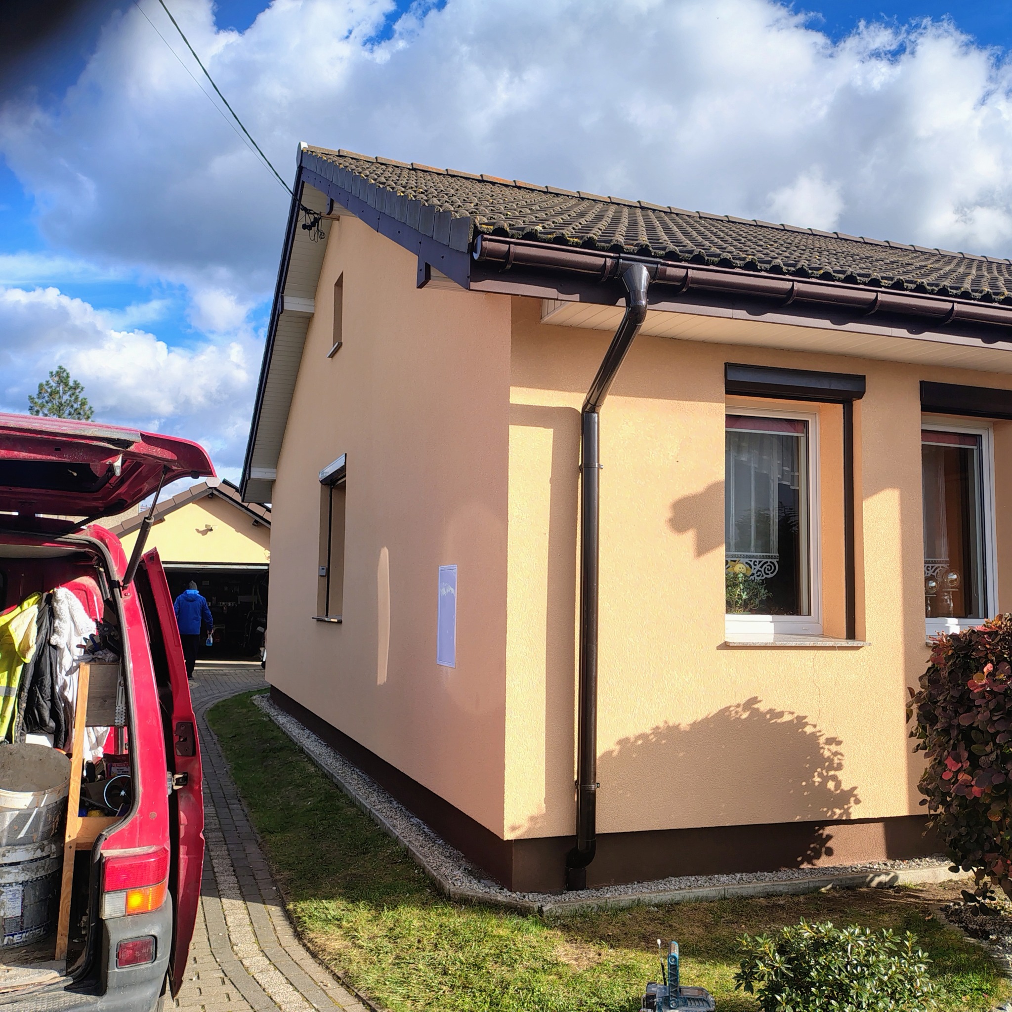 Dom jednorodzinny po renowacji elewacji w odcieniach beżu i brązu, z widocznym samochodem dostawczym i narzędziami, w słoneczny dzień.