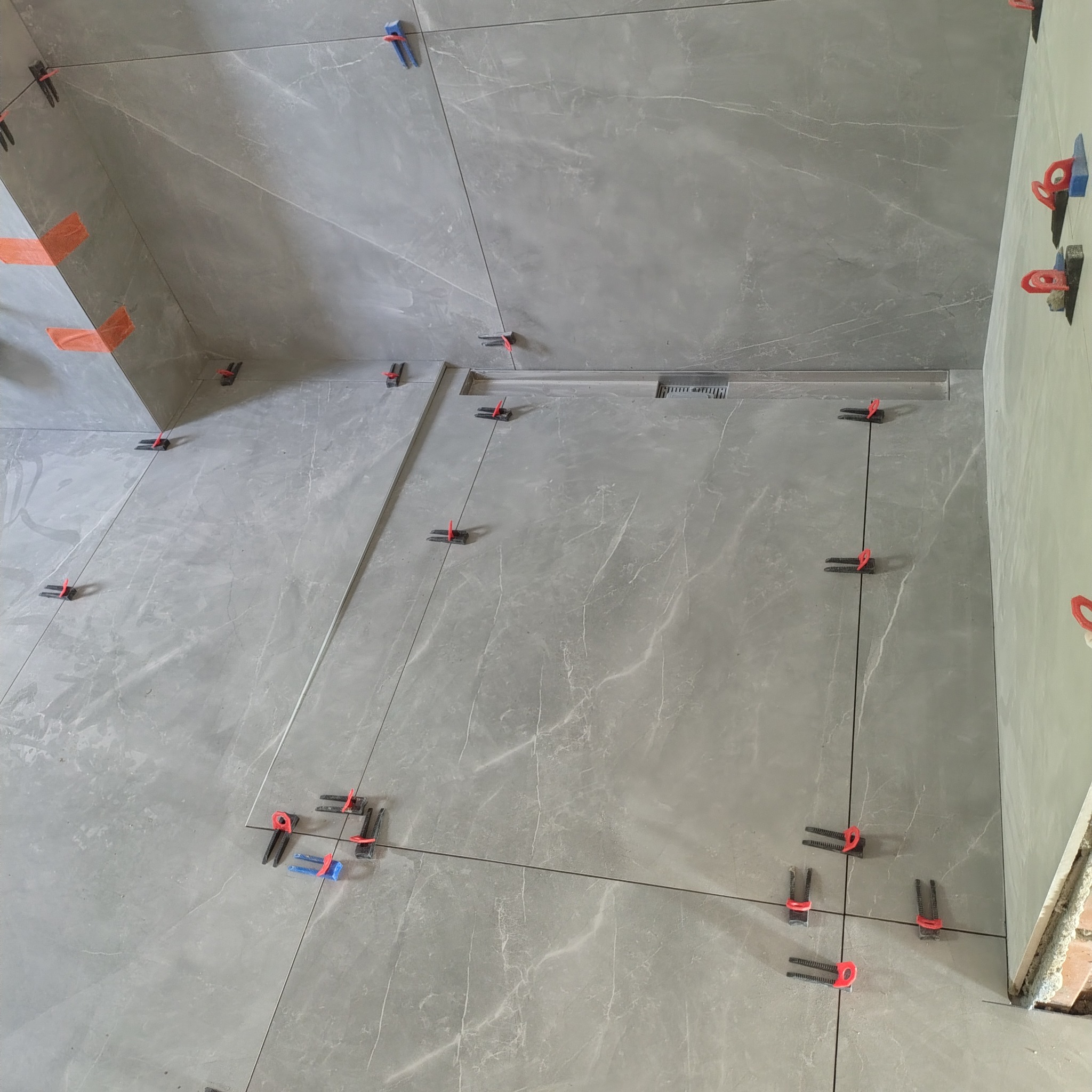 Szare płytki imitujące marmur, ułożone w łazience z użyciem systemu poziomowania. Widoczny odpływ liniowy i fragment ściany z cegły.