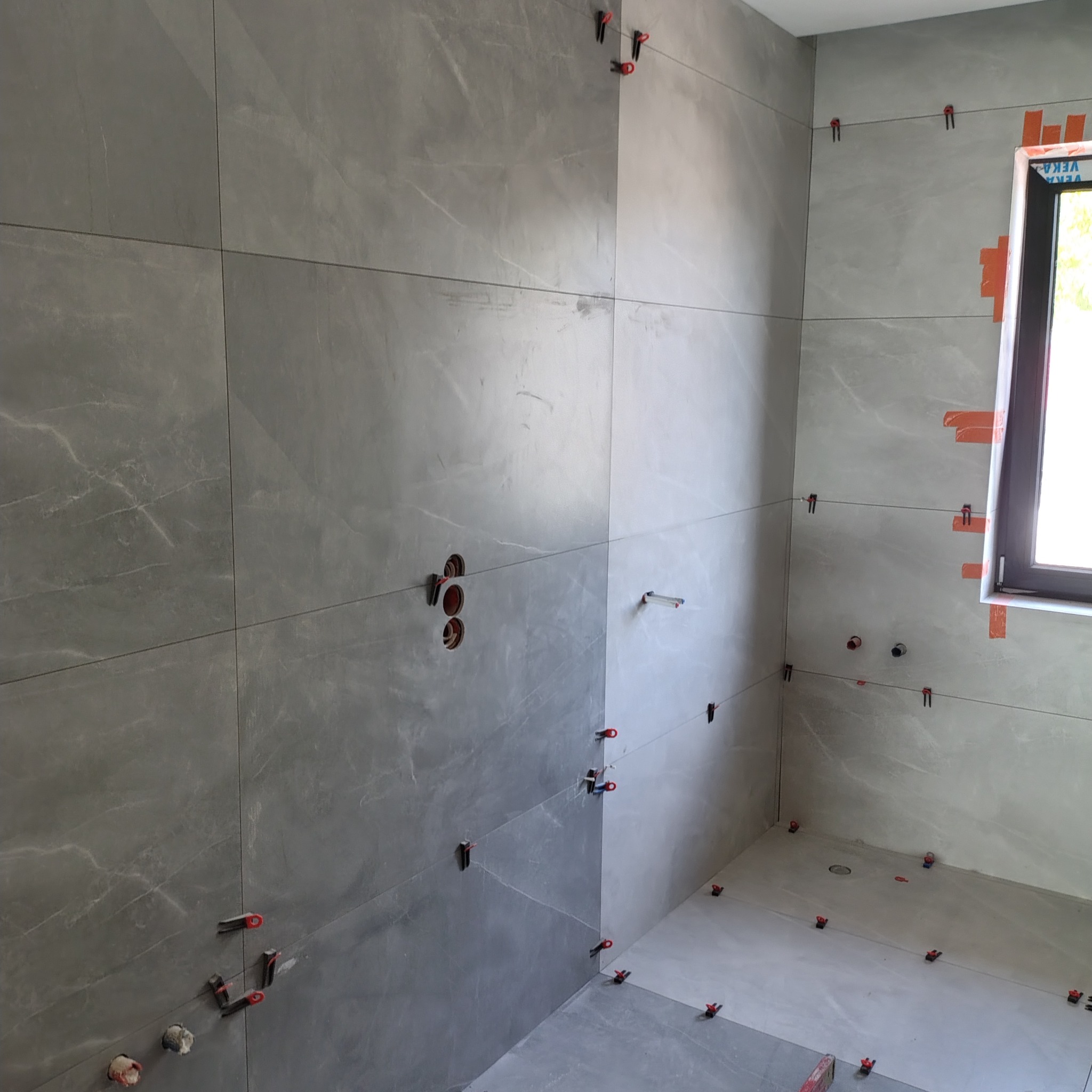 Ściany i podłoga w łazience wyłożone szarymi płytkami imitującymi marmur, z widocznymi systemami poziomowania płytek i otworami na instalacje.