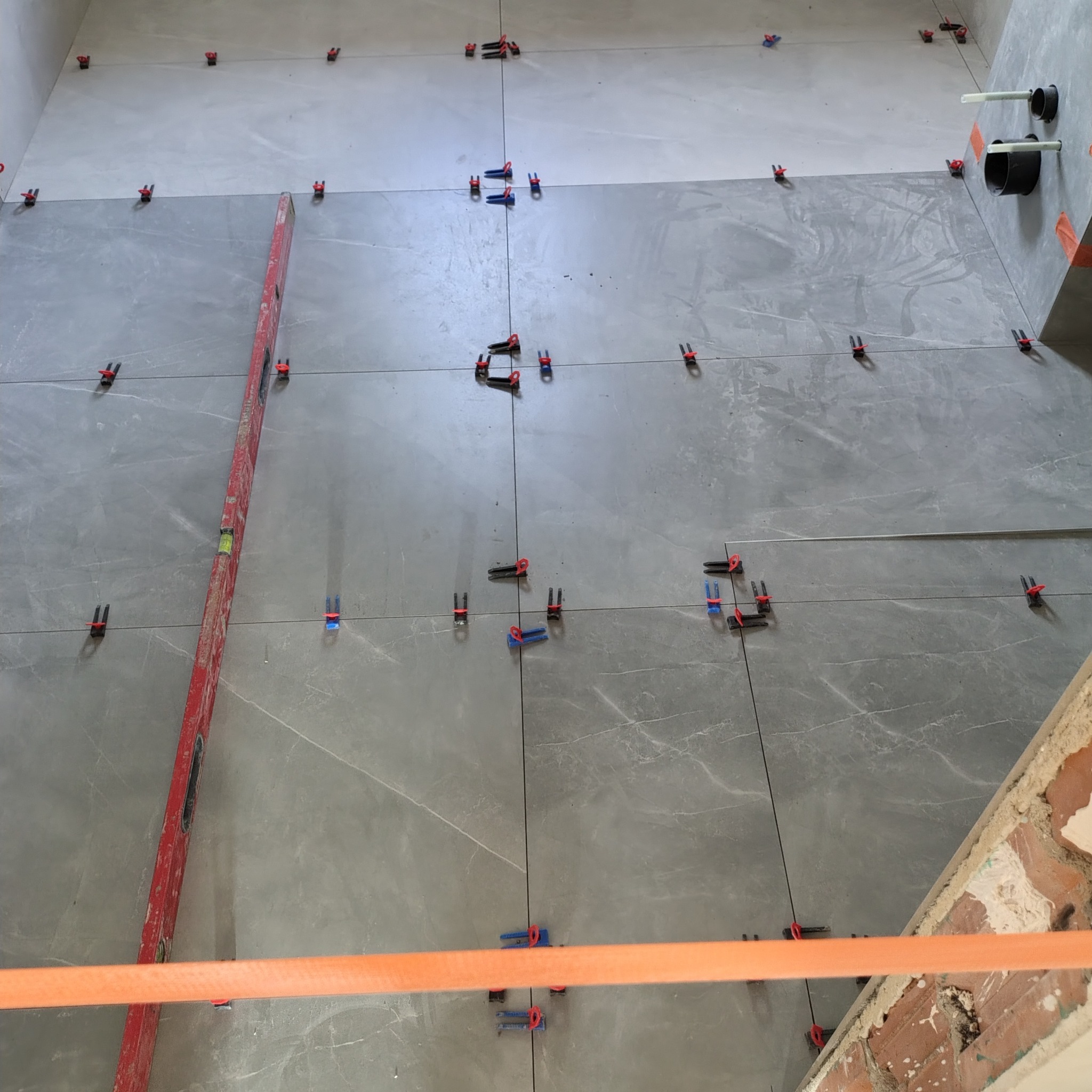 Szare płytki podłogowe w trakcie układania, z użyciem poziomicy i klipsów poziomujących. Widoczne fragmenty ściany z instalacją i surowej cegły.