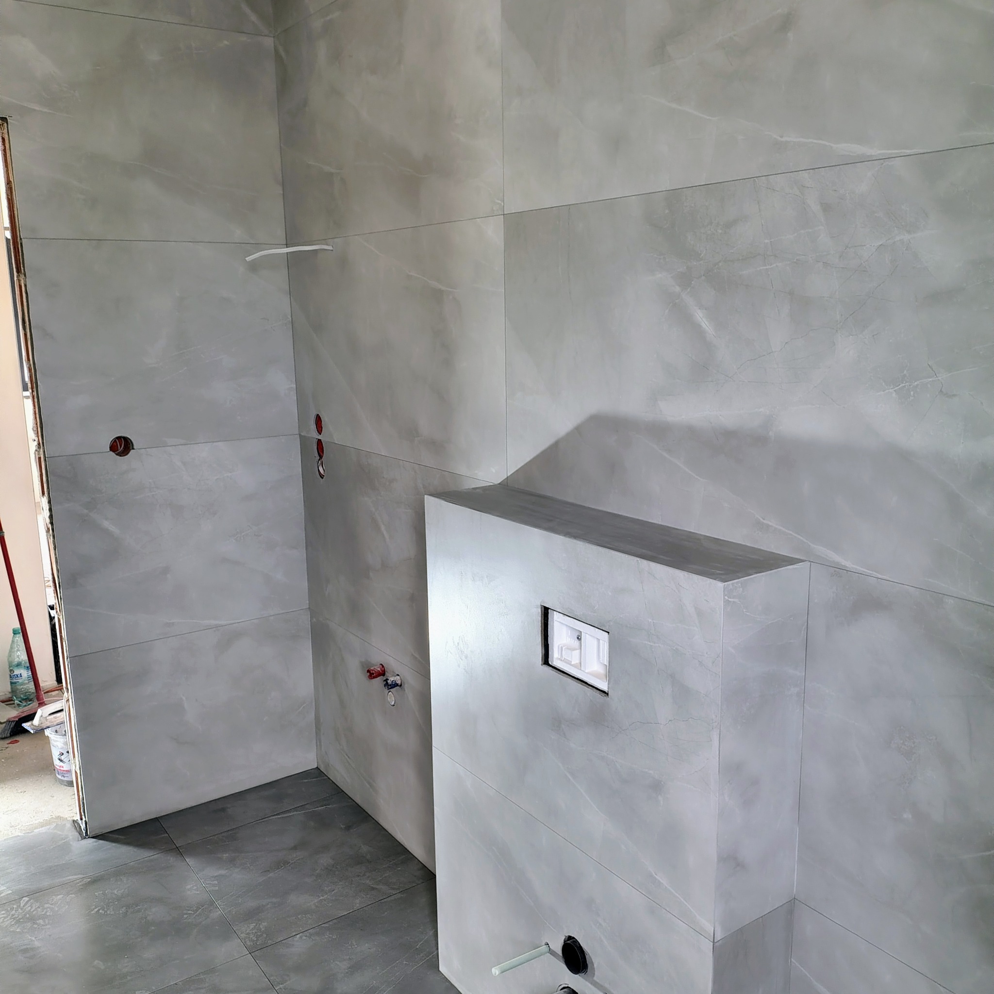 Wykończona łazienka z szarymi płytkami imitującymi marmur na ścianach i podłodze. Widoczna zabudowa stelaża WC. Minimalistyczny design, surowe wykończenie.