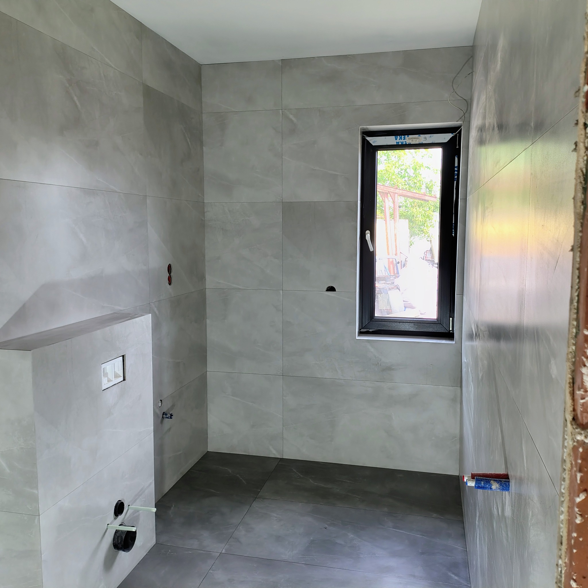 Wykończona łazienka z szarymi płytkami imitującymi marmur na ścianach i podłodze. Widoczne okno z czarną ramą i fragment instalacji sanitarnej.