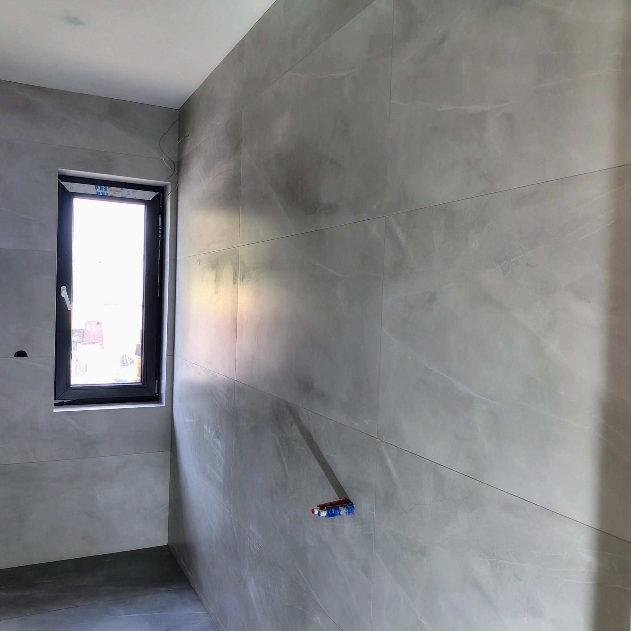 Szare, marmurowe płytki ścienne w łazience z oknem. Widoczne wystające rury instalacji wodnej. Minimalistyczny design, nowoczesne wykończenie wnętrza.