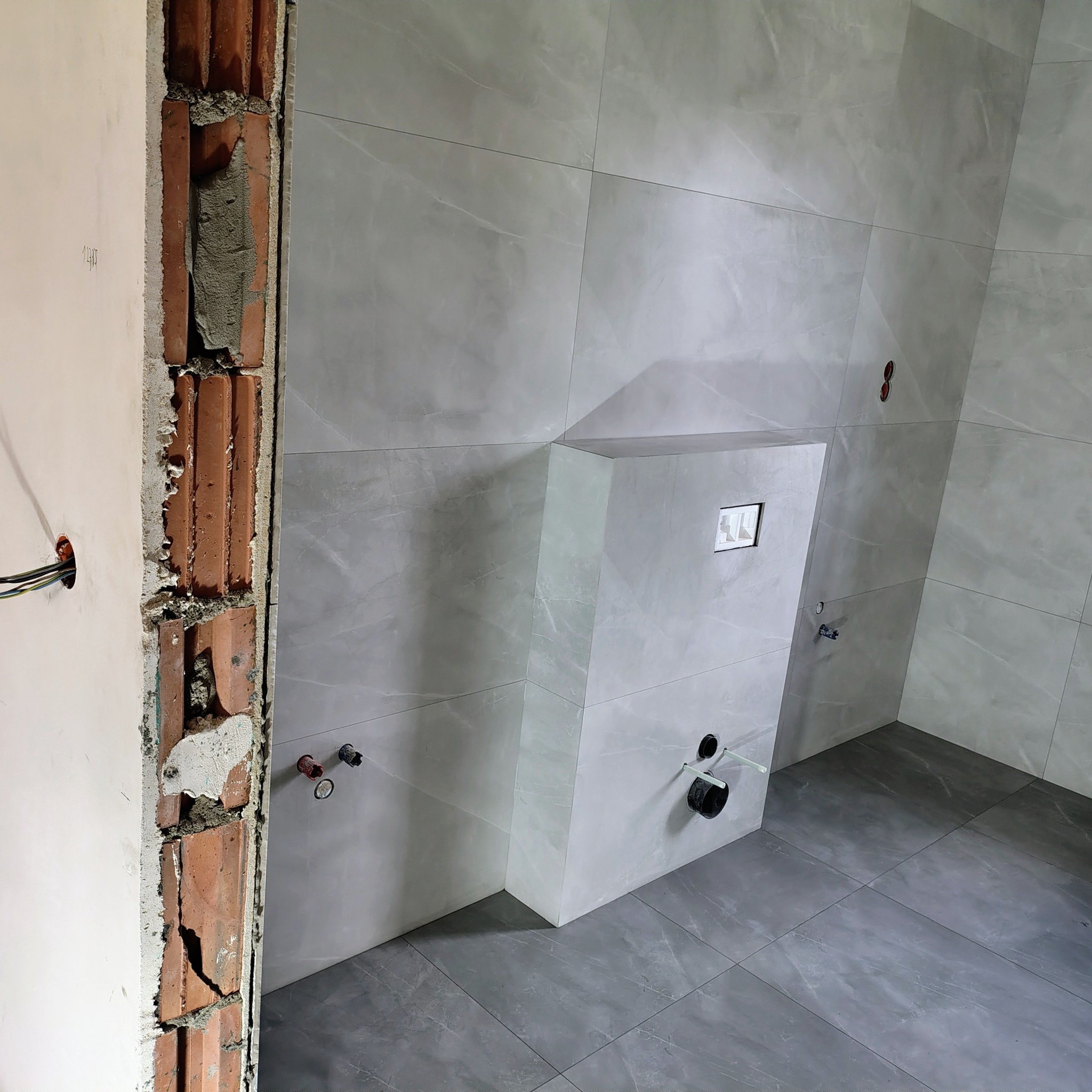 Szare płytki imitujące marmur w łazience w trakcie remontu. Widoczne instalacje wodne i elektryczne, surowa ściana z cegły. Minimalistyczny styl.