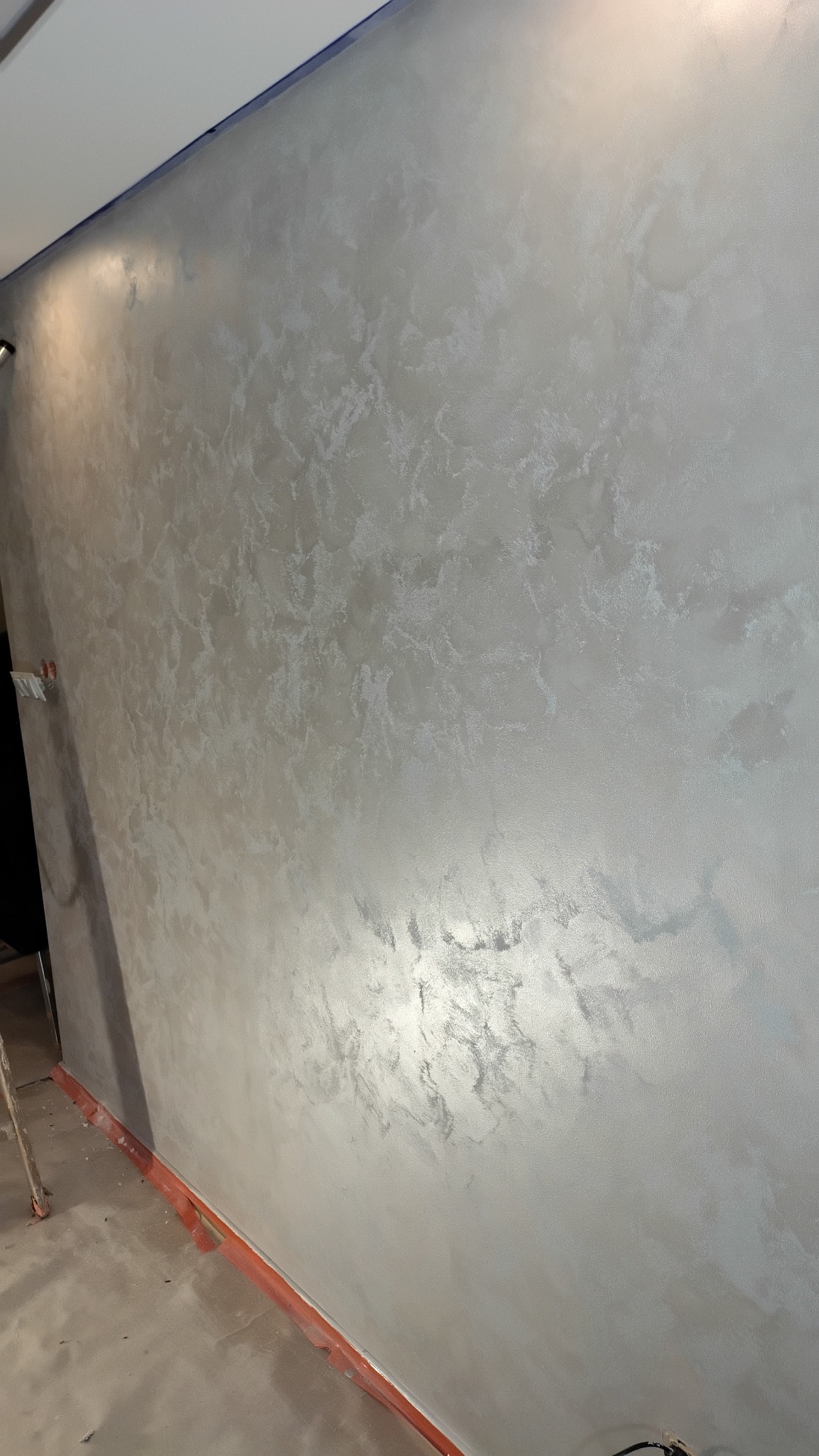 Ściana z efektem dekoracyjnym w odcieniach szarości i srebra, widoczna z bliska, z delikatnym połyskiem i nieregularną fakturą. U dołu widoczne zabezpieczenia podłogi.