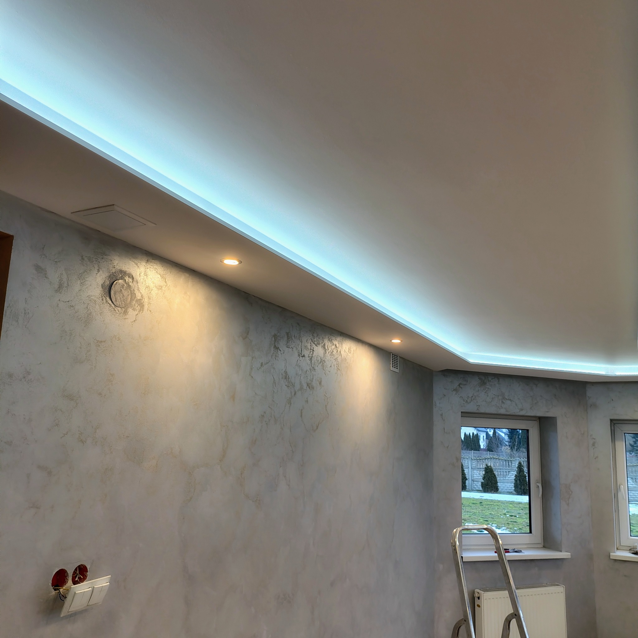 Wykończone wnętrze w Gorzowie Śląskim: dekoracyjna ściana z efektem stiuku, podświetlany sufit z punktowymi lampami. Widoczne okna i drabina.