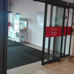 Częściowo otwarta, automatyczna szklana brama przesuwna z czerwonym napisem 'Dziękujemy, Zapraszamy ponownie', widoczna w tle automat z zabawkami.