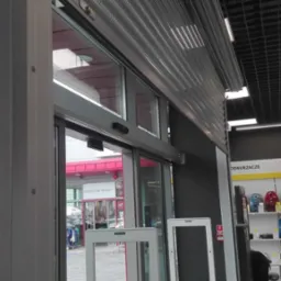 Częściowo opuszczona metalowa roleta zewnętrzna nad automatycznymi drzwiami przesuwnymi w sklepie, widoczne zabezpieczenia antykradzieżowe.