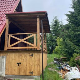 Drewport - Zadaszenia tarasów, pergole - Drewniana altana z czerwonym dachem w Bielsku-Białej, widok z zewnątrz, z elementami dekoracyjnymi w postaci czarnych zawiasów na dolnej części ścianki i krzyżowych balustrad. W tle widoczne drzewa...