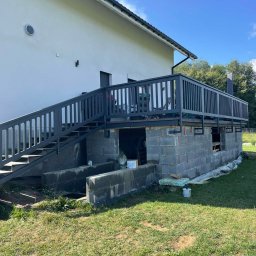 Drewport - Zadaszenia tarasów, pergole - Wznoszenie tarasu z szarą balustradą i schodami, widok na surową konstrukcję z bloczków betonowych, farba i materiały budowlane w pobliżu, dom w tle.