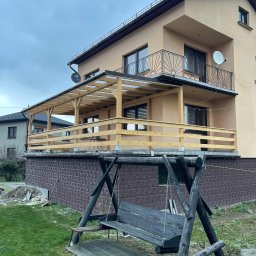 Drewport - Zadaszenia tarasów, pergole - Dwupiętrowy dom z beżową elewacją, balkonem z czarną balustradą i drewnianym tarasem na podwyższeniu z widoczną konstrukcją zadaszenia, w tle sąsiedni dom i pochmurne niebo, na pierwszym planie...