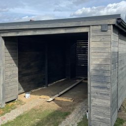 Drewport - Zadaszenia tarasów, pergole - Nowoczesna, otwarta konstrukcja z szarego drewna z płaskim dachem i widocznymi śladami impregnacji, częściowo wykończona, z widocznymi deskami i wiadrem farby w środku.