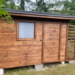 Drewniana altana ogrodowa z oknem z pleksi i kratką, podniesiona na betonowych bloczkach, otoczona zielenią.