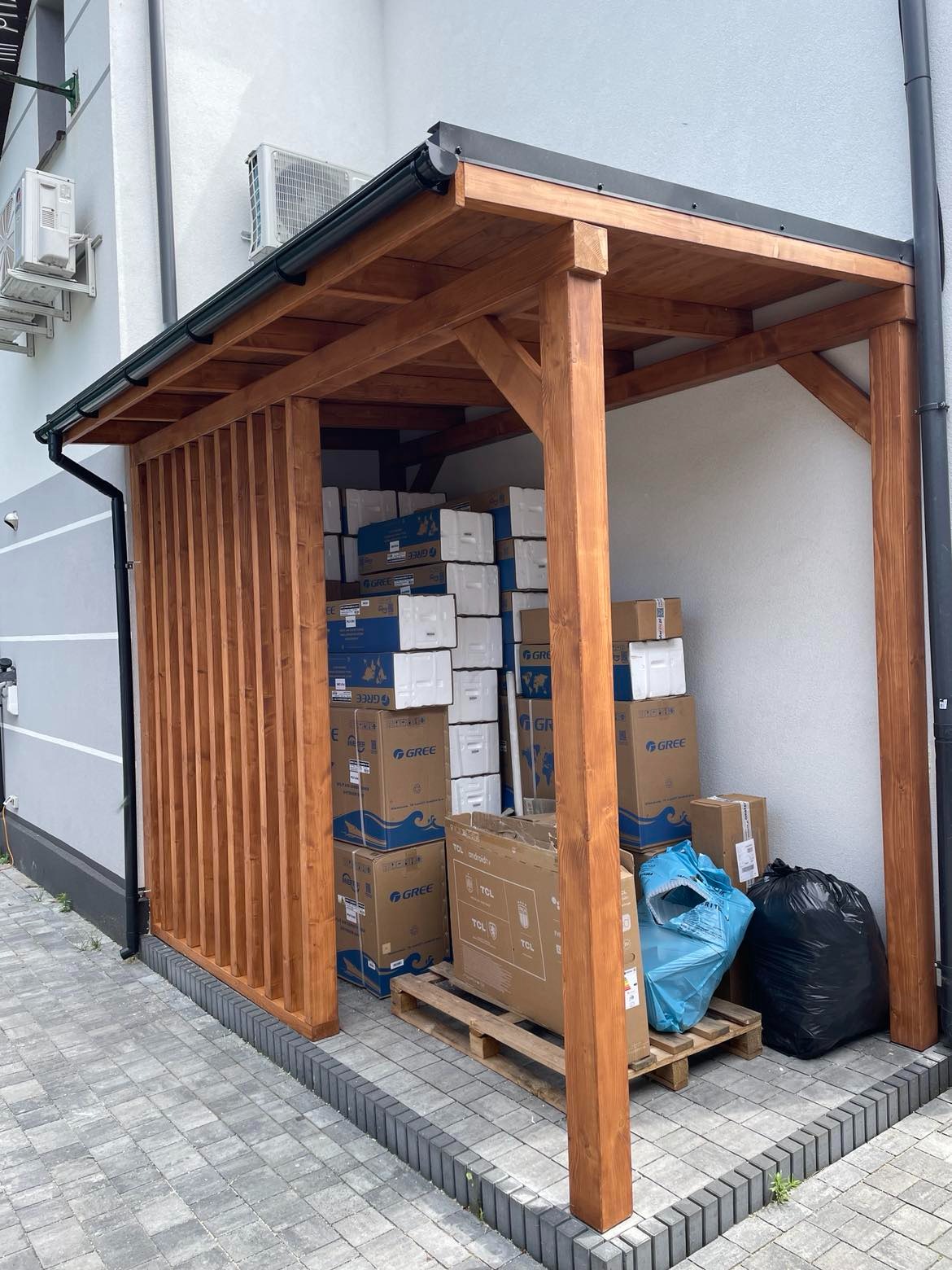 Drewniana konstrukcja zadaszenia przy ścianie budynku, wypełniona kartonami z klimatyzatorami Gree i telewizorami TCL, stojąca na palecie, z workami na śmieci obok.