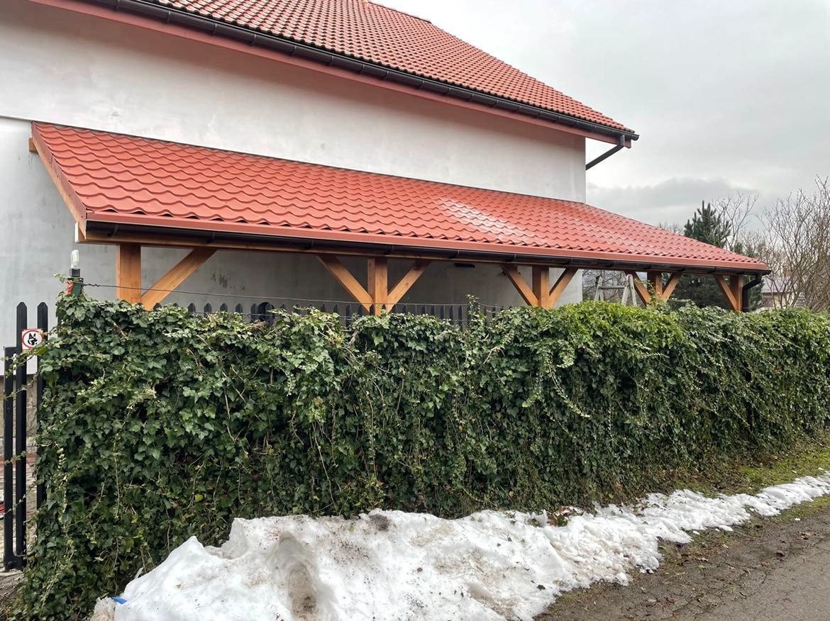 Zadaszenie tarasu z czerwoną dachówką wsparte na drewnianych belkach, częściowo zasłonięte przez gęsty żywopłot z bluszczu i leżący śnieg.