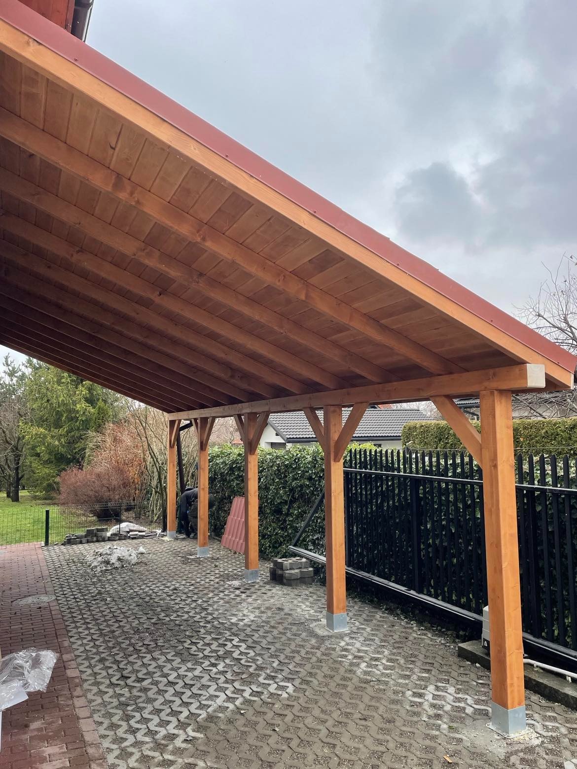 Drewniana konstrukcja z czerwonym dachem na posesji, widoczna w trakcie montażu, z elementami wciąż do ułożenia na brukowanym podłożu i pracującą osobą w tle.