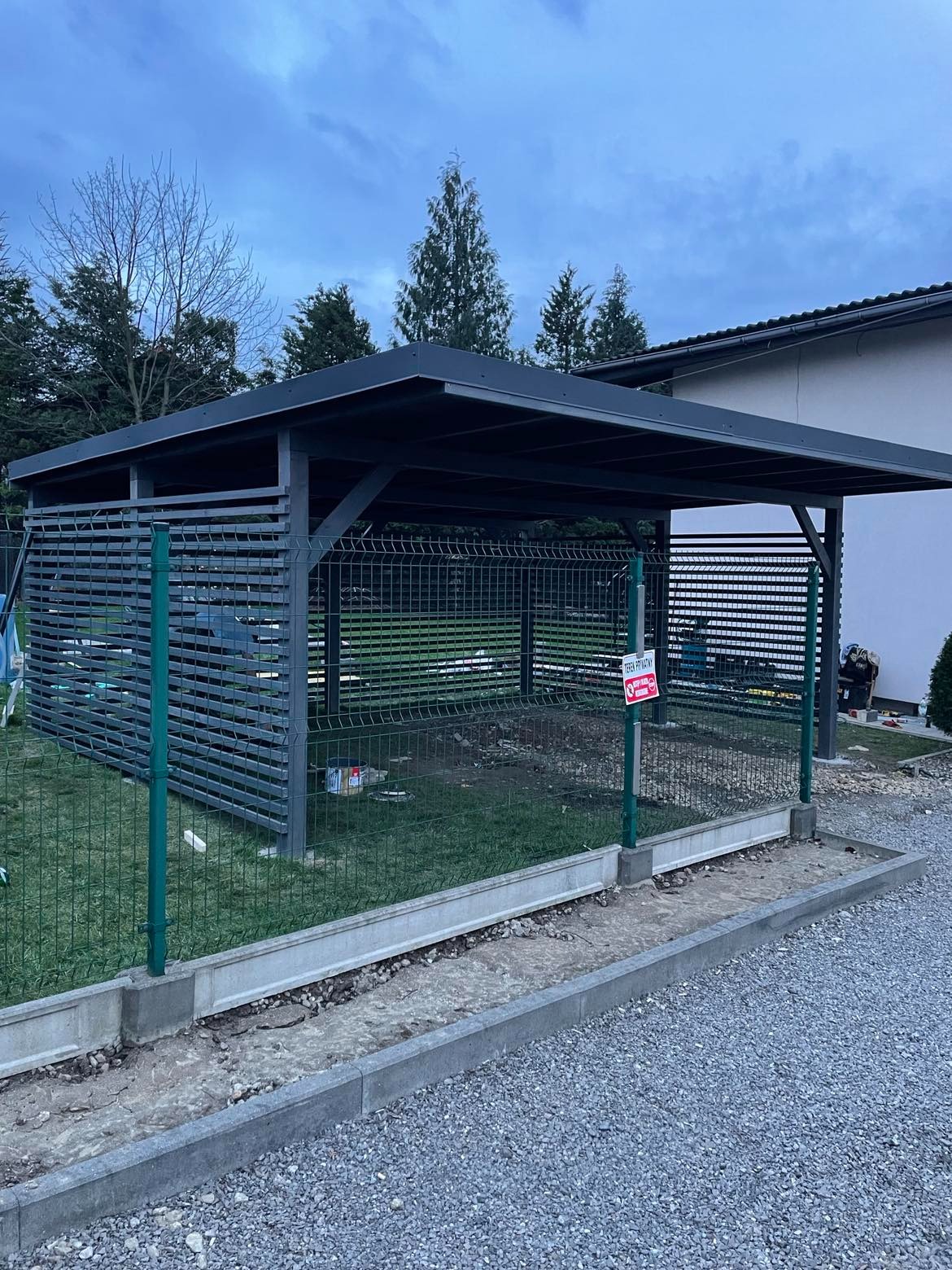 Nowoczesna, grafitowa pergola z metalową konstrukcją i ażurowymi ścianami, otoczona zielonym ogrodzeniem i świeżo ułożonym obrzeżem z kostki, na tle domu i drzew.