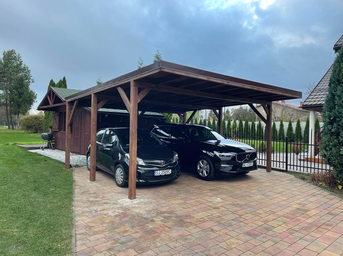 Zadaszenie drewniane w kolorze ciemnego brązu, pod którym zaparkowane są dwa czarne samochody osobowe na posesji z kostki brukowej. W tle widoczny domek narzędziowy i zadbany ogród z tujami.