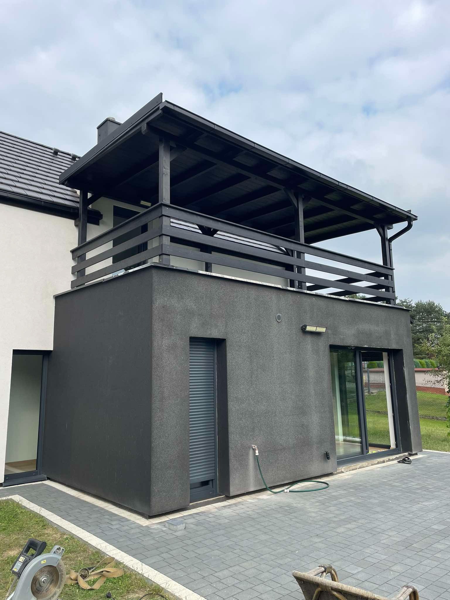 Nowoczesny dom z płaskim dachem i czarną drewnianą pergolą na balkonie, szare ściany i kostka brukowa na podwórku, widoczne narzędzia budowlane.