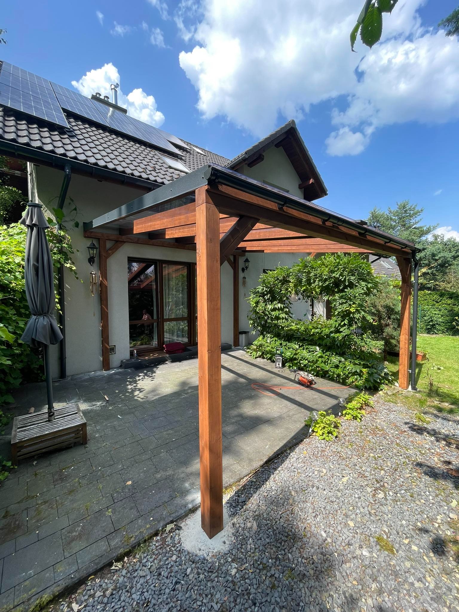 Drewniana pergola z przezroczystym zadaszeniem przy domu z panelami słonecznymi, widok na taras z szarą kostką brukową i roślinność.