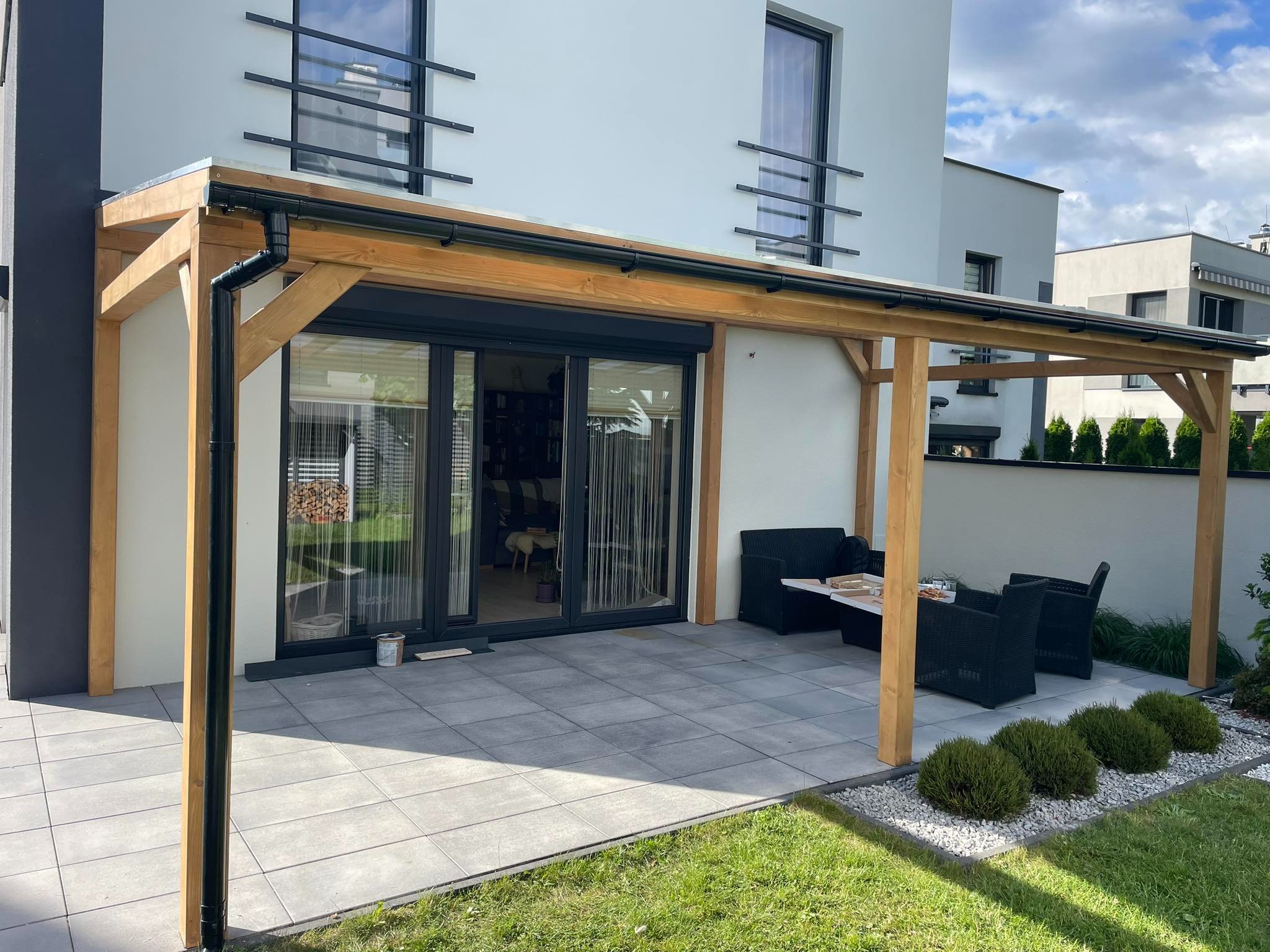 Zadaszona pergola drewniana z czarnymi rynnami, przylegająca do nowoczesnego domu z szarymi płytami tarasowymi i czarnymi meblami ogrodowymi.