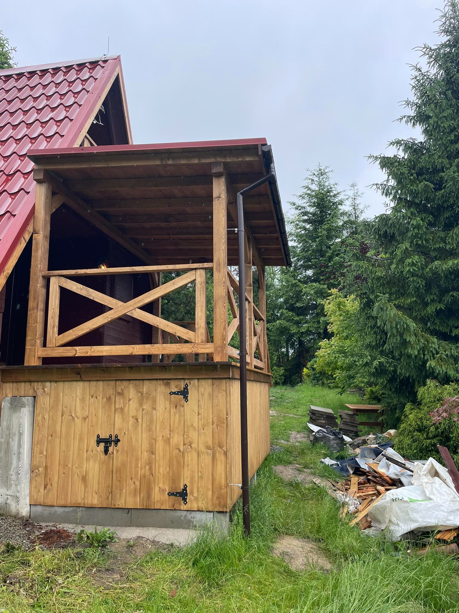 Drewniana altana z czerwonym dachem w Bielsku-Białej, widok z zewnątrz, z elementami dekoracyjnymi w postaci czarnych zawiasów na dolnej części ścianki i krzyżowych balustrad. W tle widoczne drzewa...